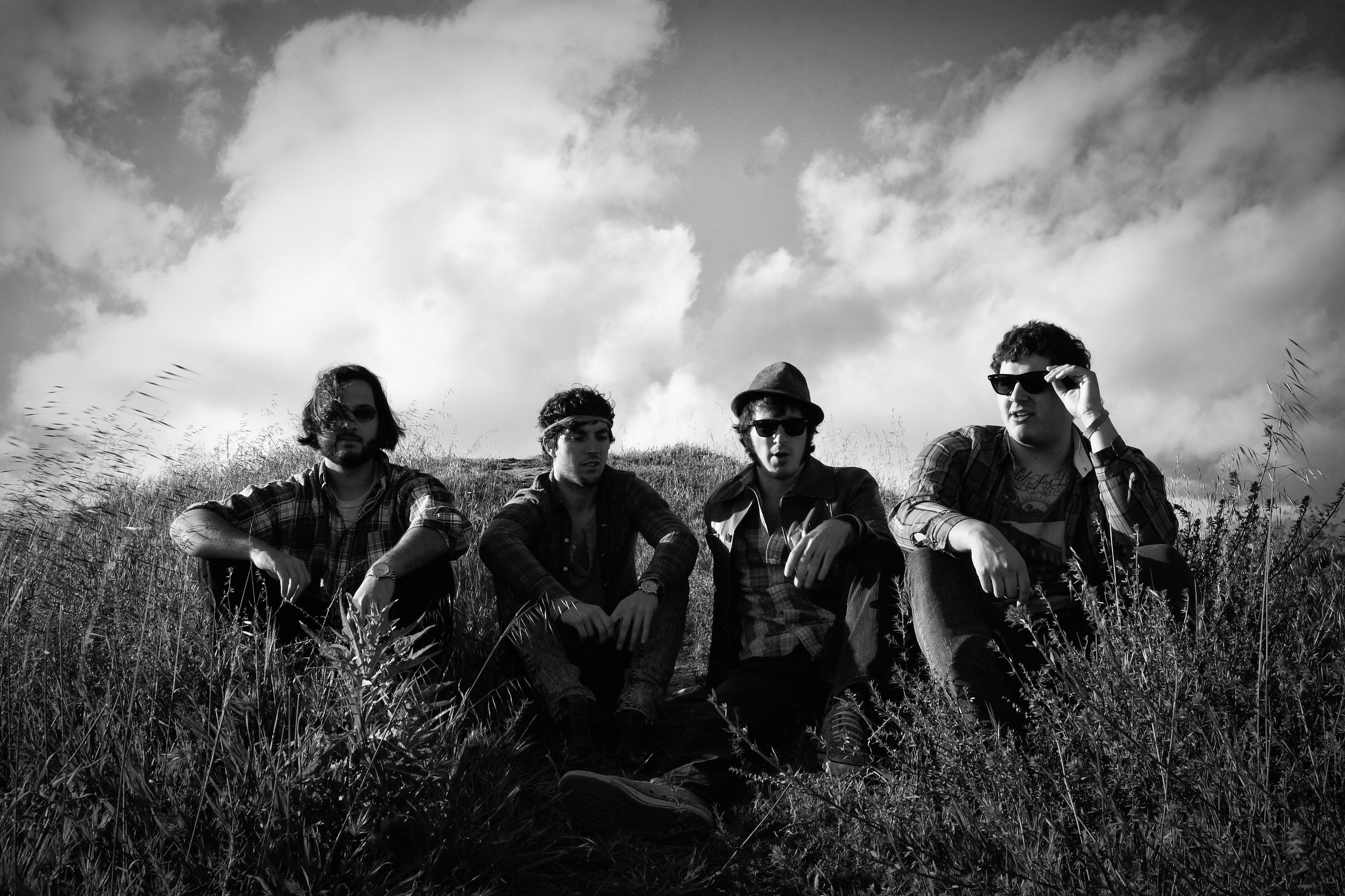 Black and white photo of four young men sitting on grass outdoors under a partly cloudy sky, wearing sunglasses and casual clothing.