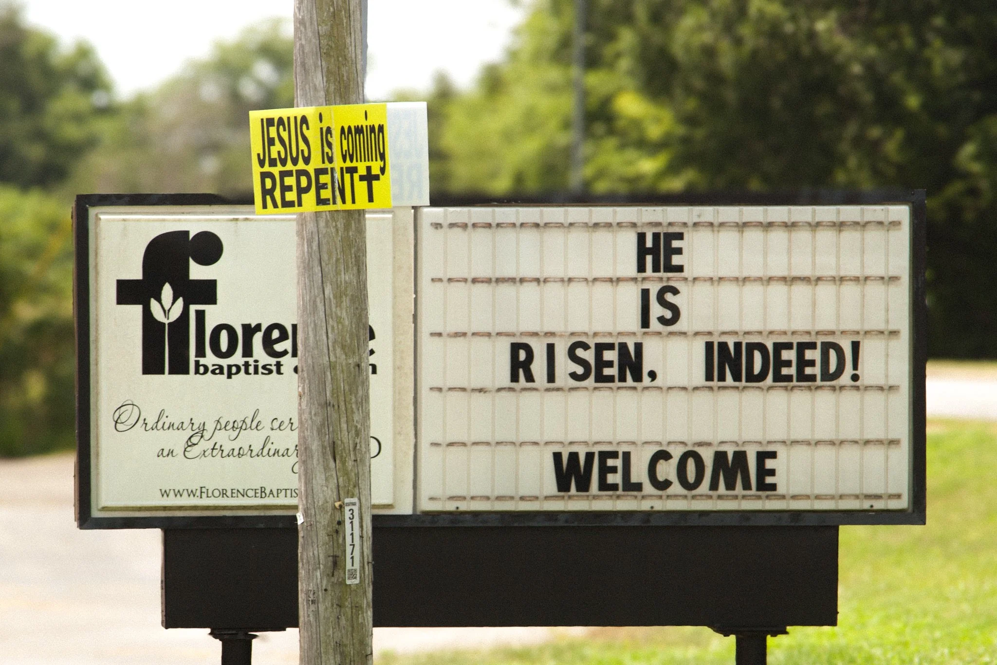 A church sign with a Christian message and a yellow sticker that reads "JESUS is coming REPEAT" attached to a wooden pole.