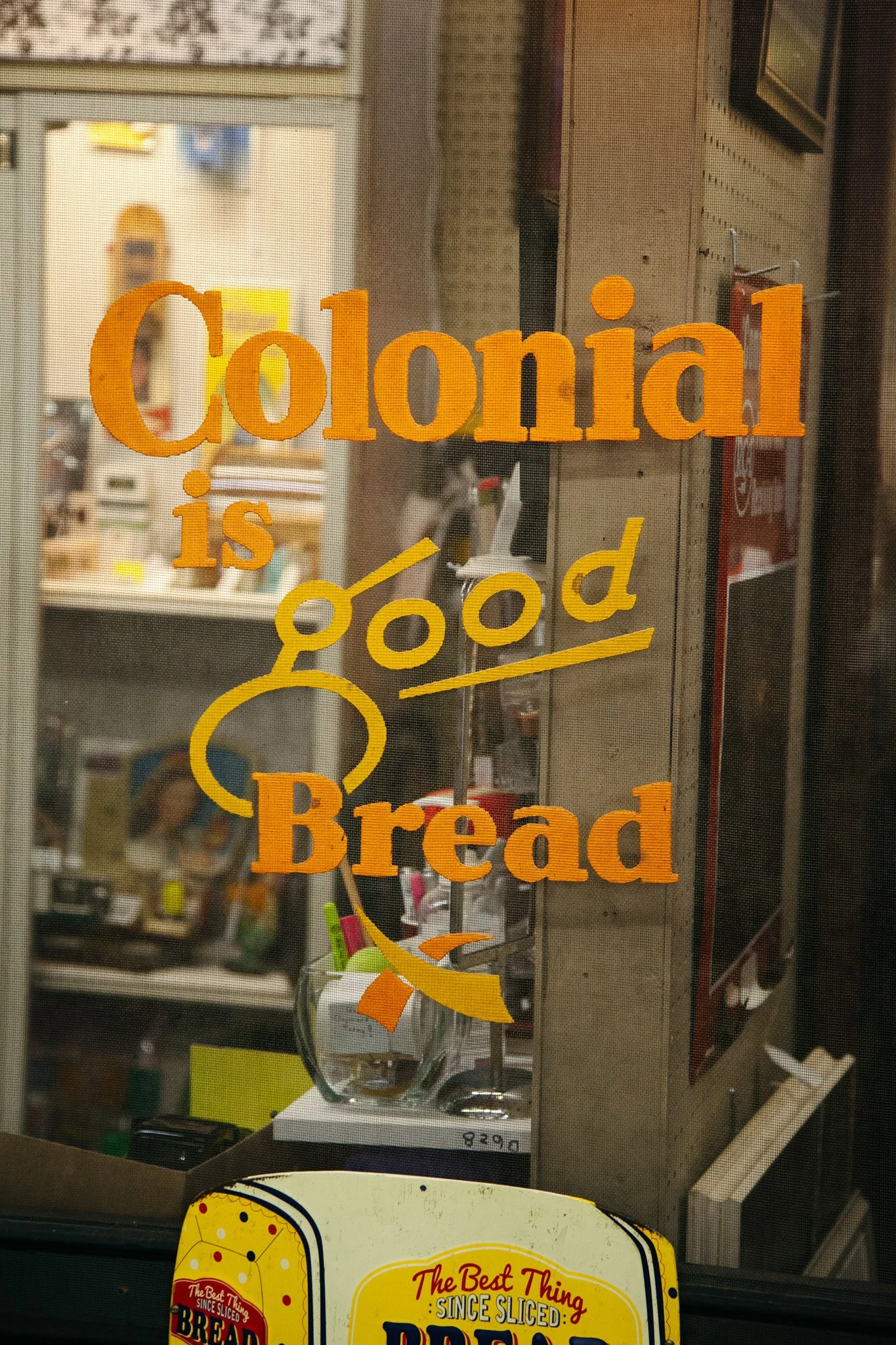 Close-up of a glass door with an orange and yellow sign that reads 'Colonial is good Bread'. The interior of a grocery store with shelves and products visible through the glass.