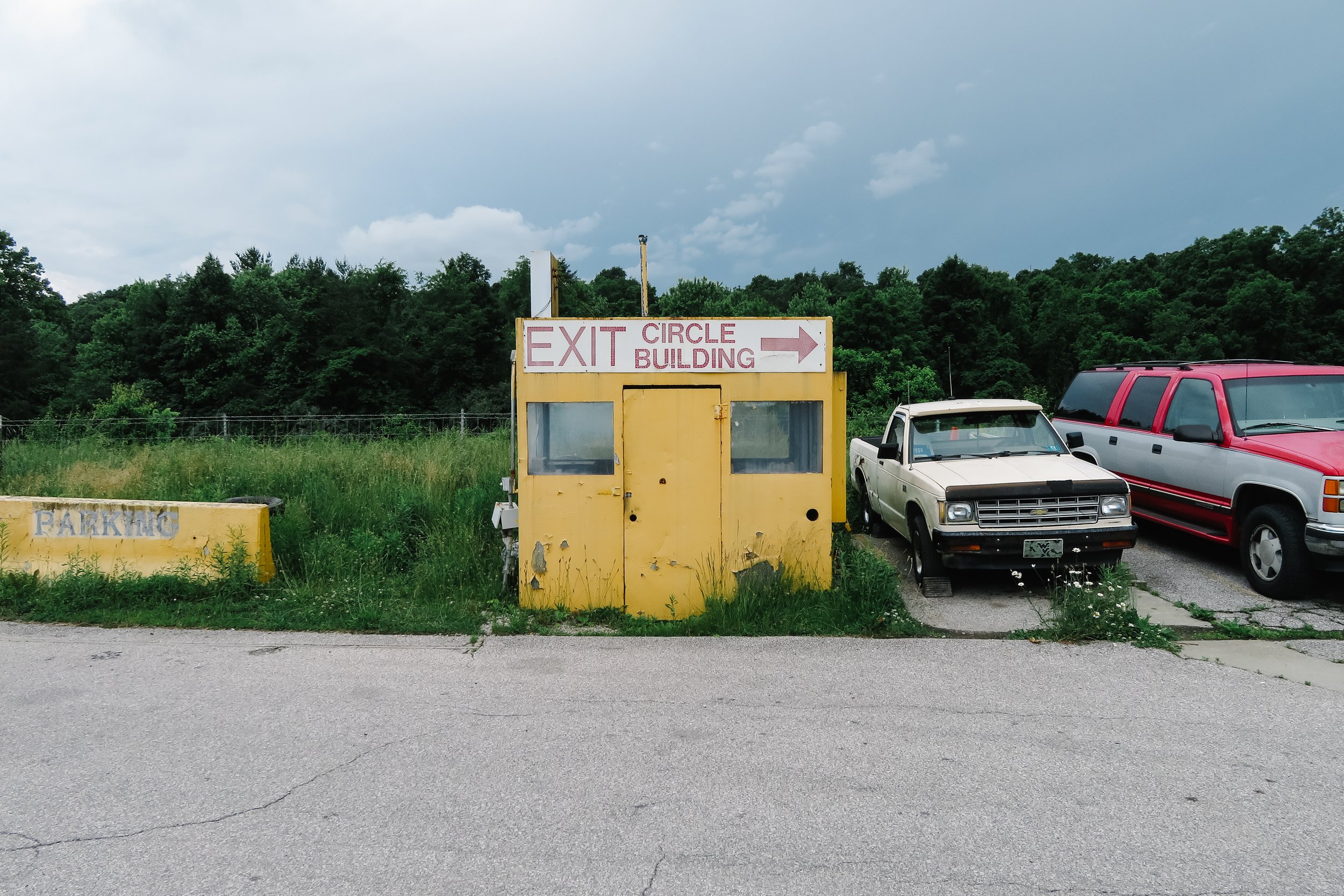 A parking lot with a yellow exit sign and a small white truck parked next to a red vehicle. The sign indicates the exit for the circle building to the right. The scene is set against a backdrop of green trees and a cloudy sky.