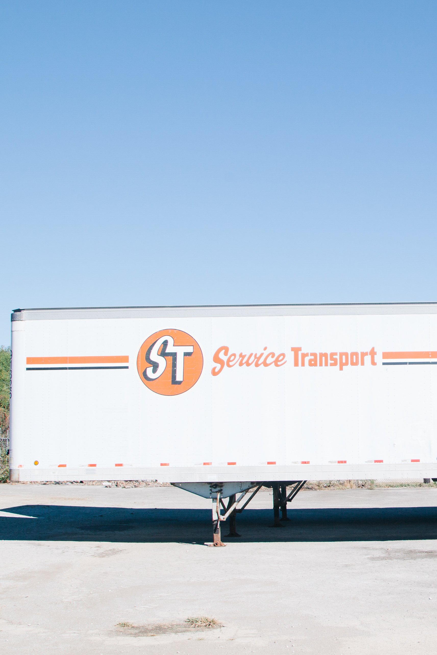 A white trailer with orange and black small stripes, displaying the logo of a company with the initials 'ST' and the words 'Service Transport' underneath. The trailer is supported by metal jacks and is parked on a concrete surface, with a clear blue 