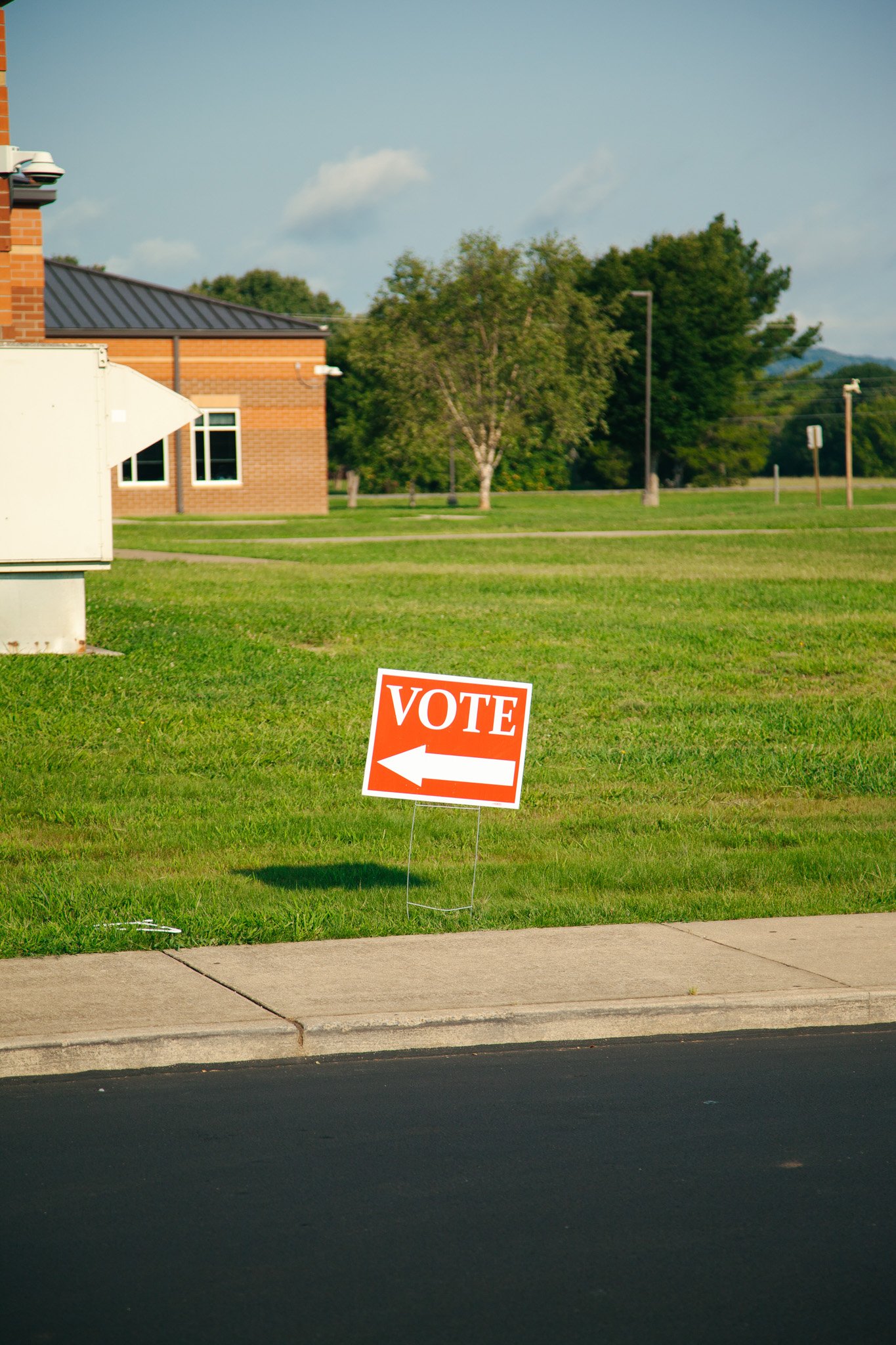 A red and white sign on the grass near a sidewalk says 'VOTE' with a left arrow. In the background, there is a brick building, a tree, and a few street lights under a blue sky.