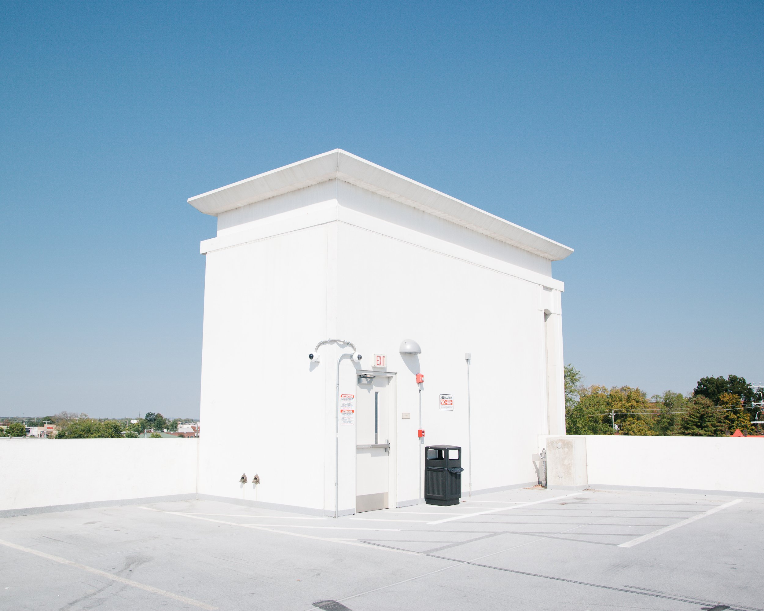 Rooftop parking lot with white building at the corner, trash bin, and multiple parking spaces, under a clear blue sky.