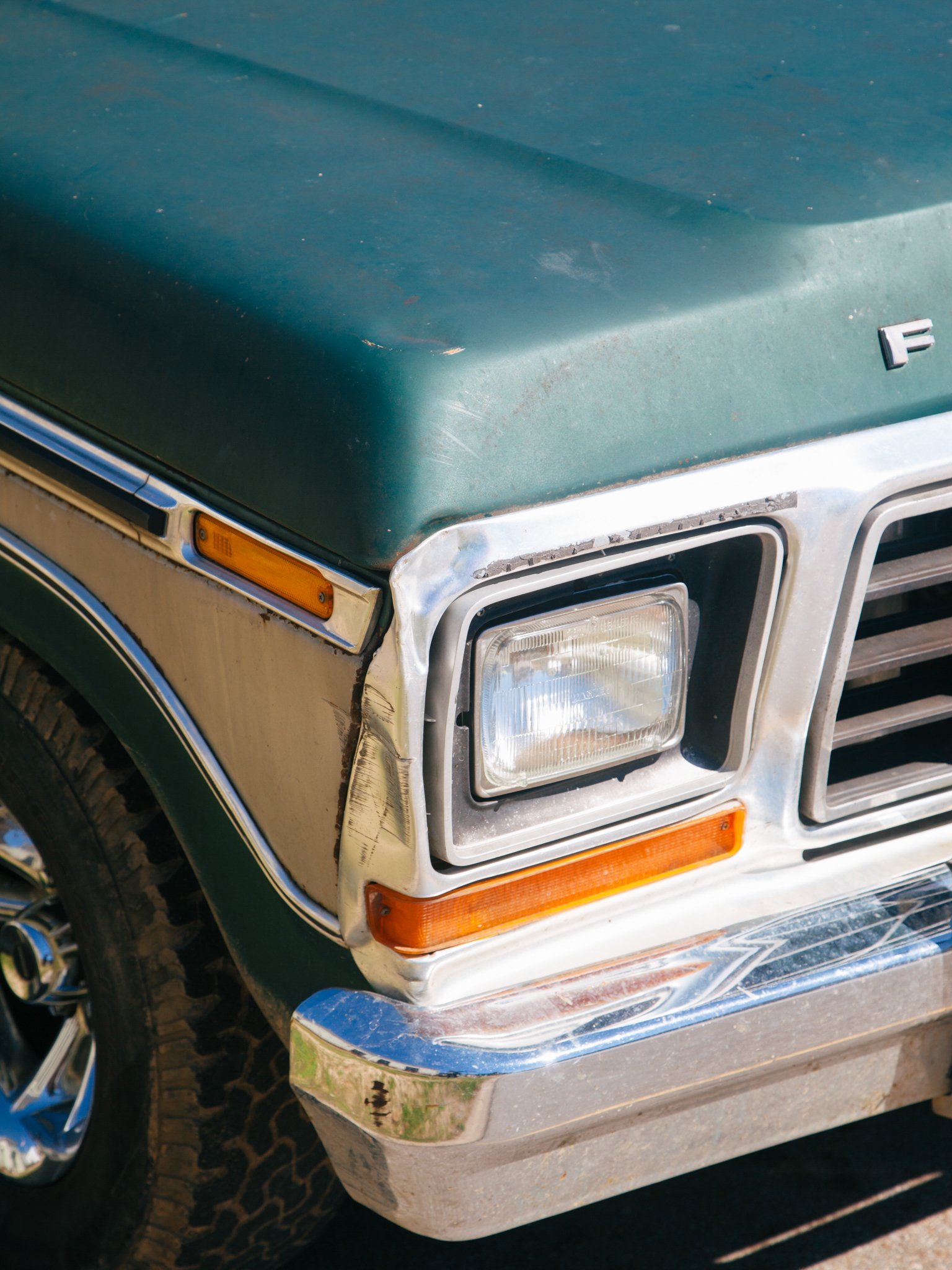 Close-up of the front right corner of a vintage green SUV with visible damage, rust, and dirt on the bumper and headlight area.