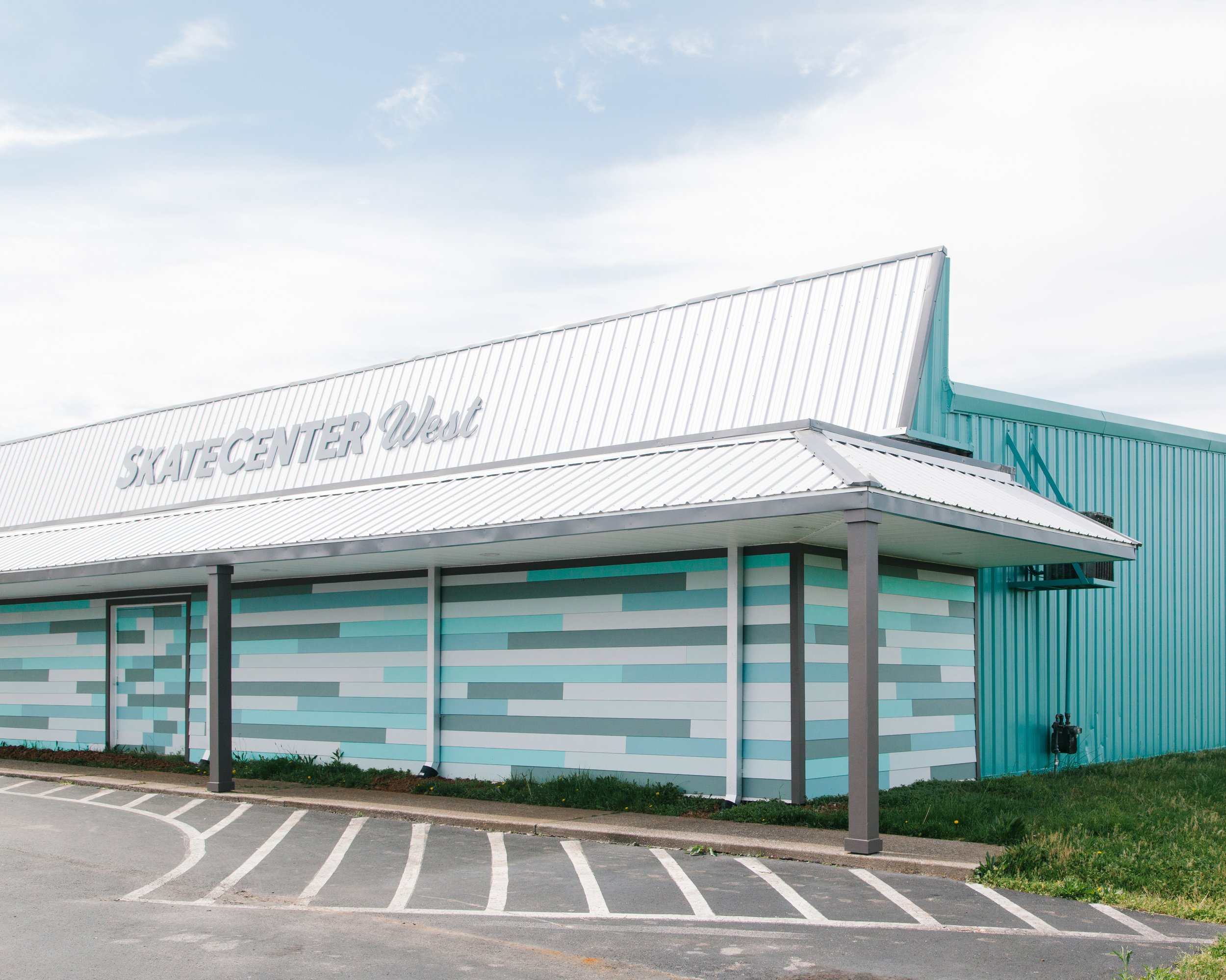 Exterior of a light blue and white sports complex building with the words 'SKATE CENTER West' on the front, adjacent parking lot with marked spaces, and a partly cloudy sky overhead.