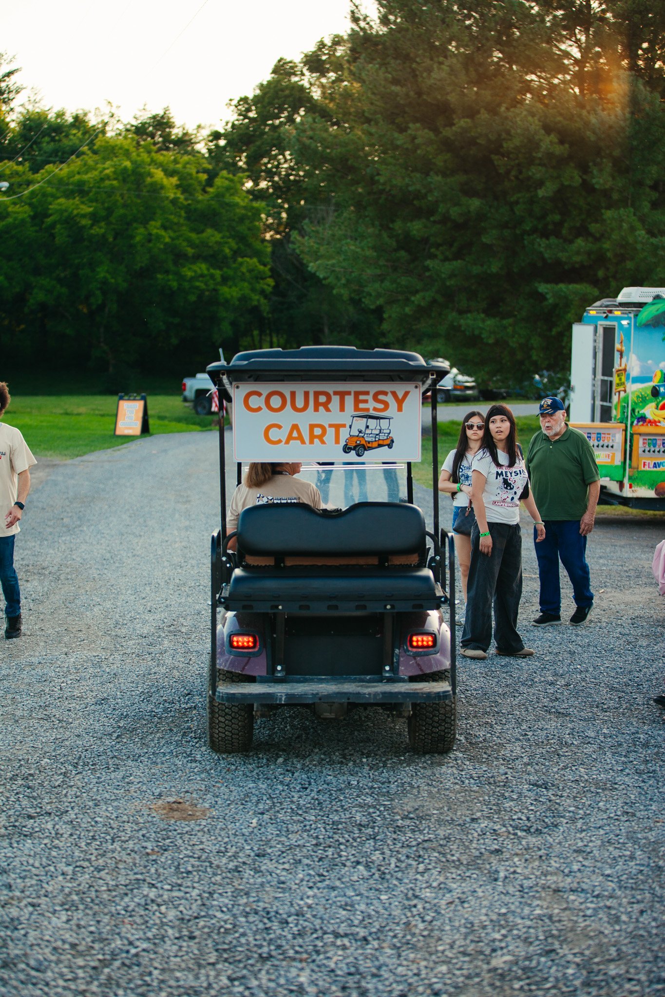People walking near a golf cart with a sign that says 'Courtesy Cart' at an outdoor event during sunset, with green trees and food trucks in the background.