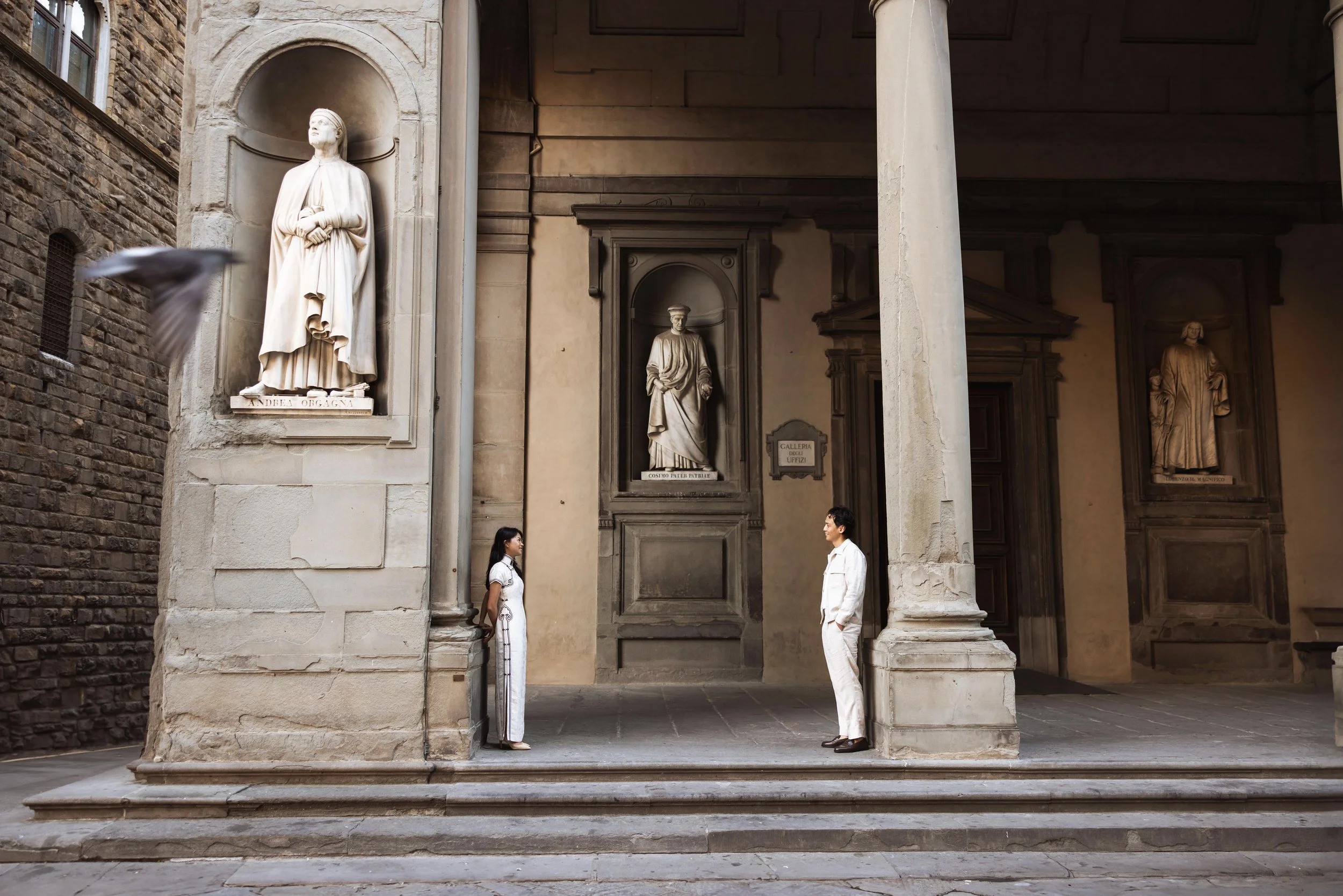 Two people standing face to face on a city sidewalk under a stone archway with statues in recessed niches behind them and a bird in flight on the left.