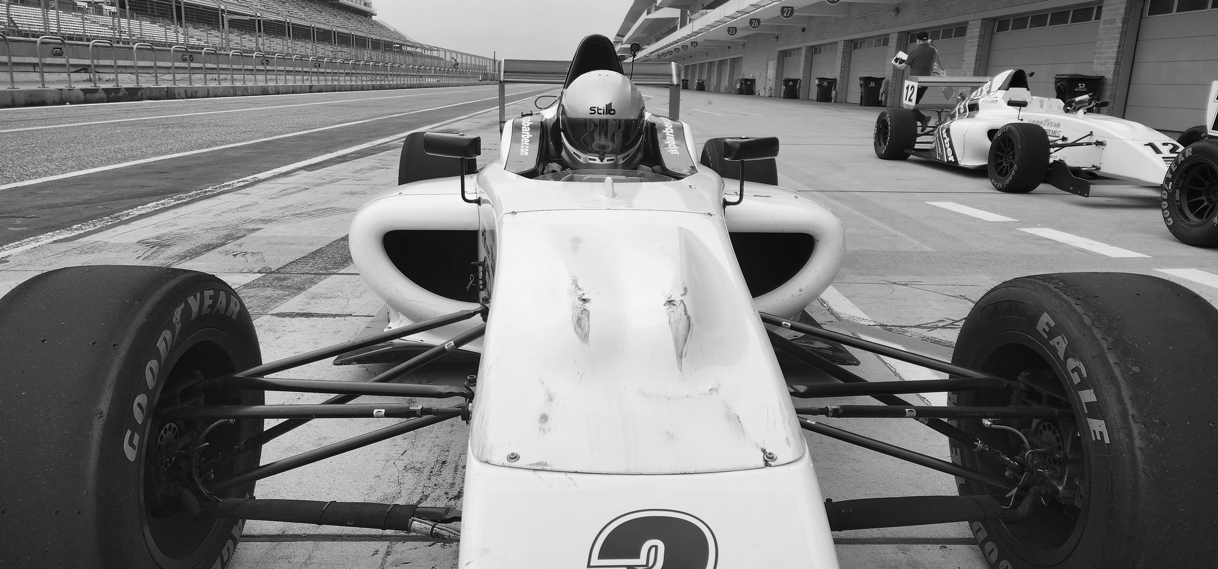 Black and white photo of race cars in a pit lane, focusing on a Formula racing car with visible Goodyear tires and a driver wearing a helmet.