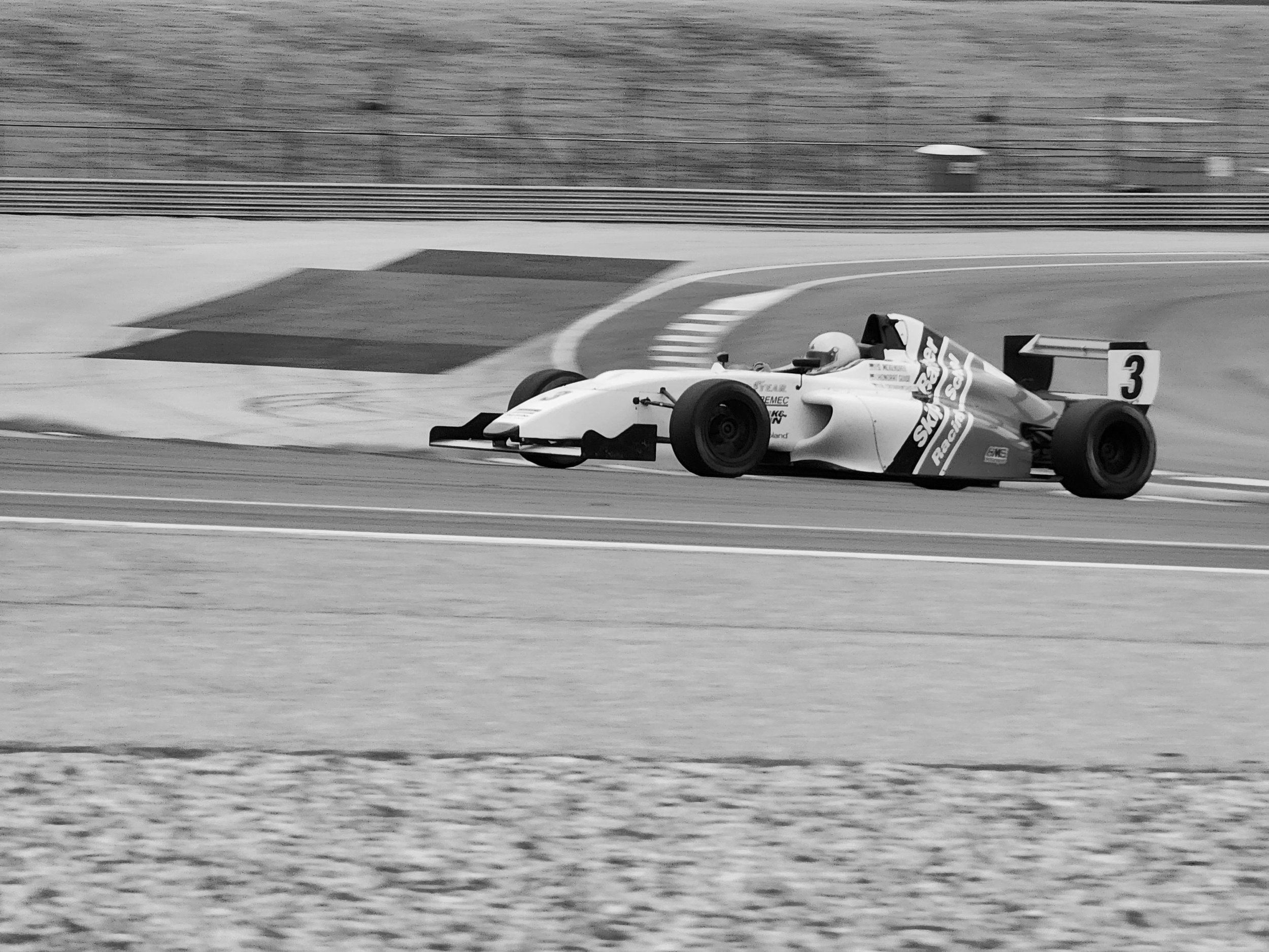 Black and white image of a Formula race car on a track, with the number 3 on its side and various sponsor logos.