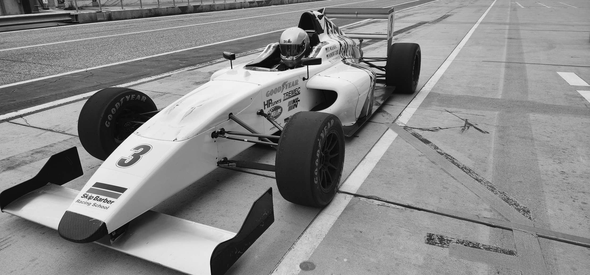 Black and white image of a single-seater race car marked with number 3, parked on a racetrack pit lane.