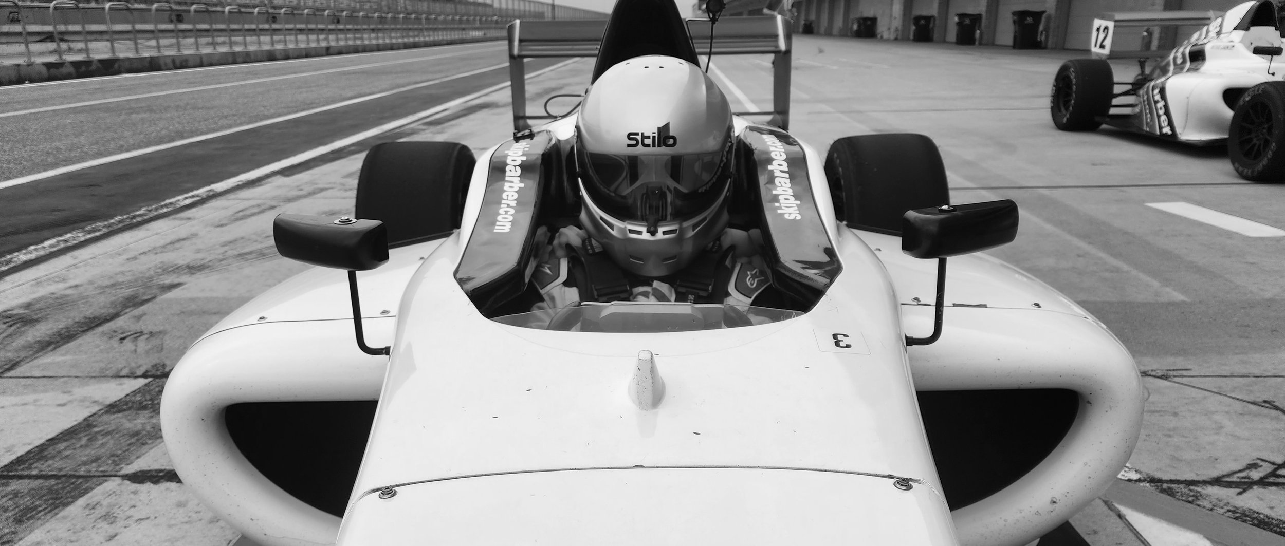 Race car with driver in helmet at a racetrack pit lane, displaying a close-up front view of the vehicle.