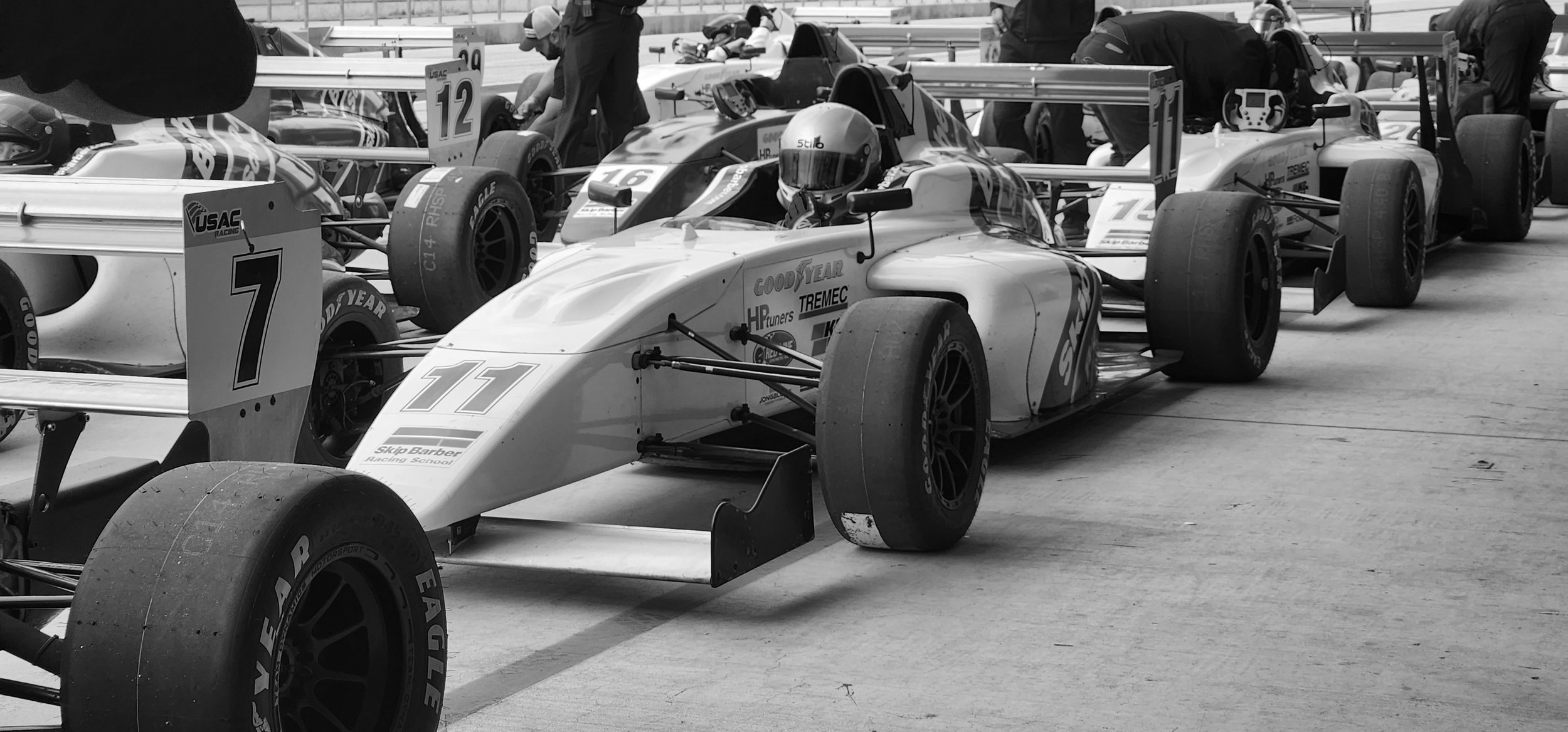 Black and white image of race cars lined up in a pit row, featuring various number and sponsor logos. Drivers in helmets are seated in the cars, and crew members are seen around, preparing for a race.