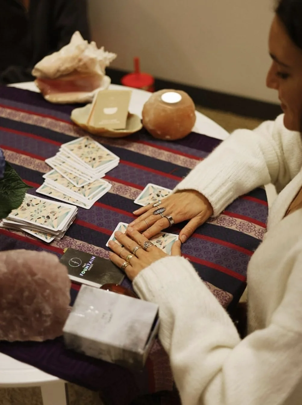 A woman is reading tarot cards at a table that is decorated with crystals, rocks, and salt lamps.