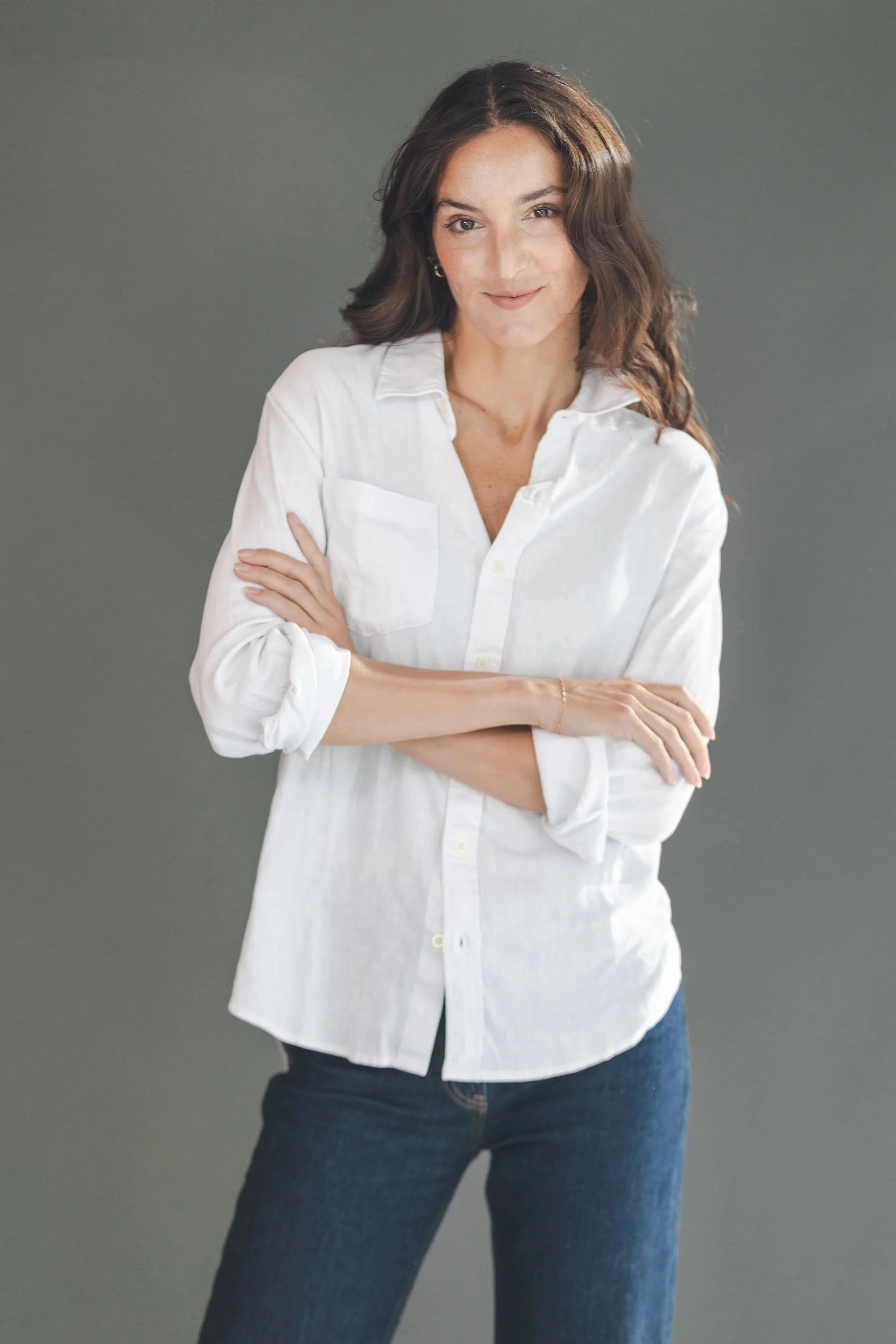 A woman with wavy brown hair wearing a white button-down shirt with sleeves rolled up and dark blue jeans, standing with arms crossed against a plain gray background.