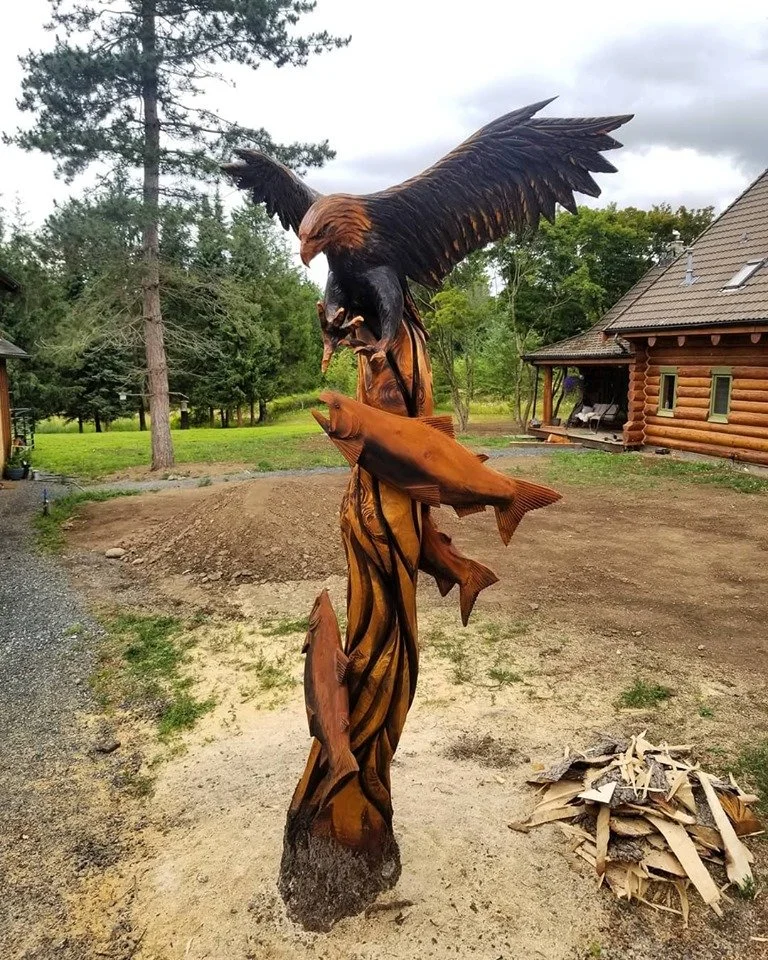 Wooden sculpture of a bald eagle with wings spread, perched on a base, with three fish carved on the sculpture's body, outdoors in front of a log cabin in the Seattle area, by local chainsaw carver Tomas Vrba