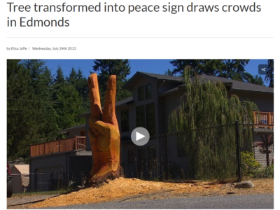 A large tree sculpted into a peace sign with two fingers raised, located in front of a house with trees in the background.