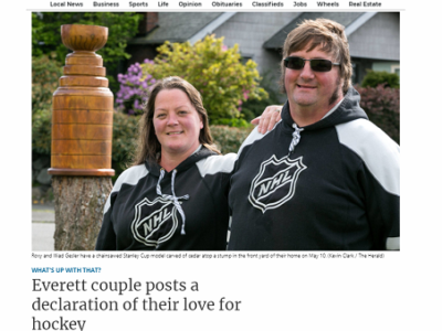 A smiling woman and man, both wearing NHL hoodies, standing outdoors in front of a chainsaw carved wood sculpture of the Stanley Cup, created by Bellingham sculptor Tomas Vrba.