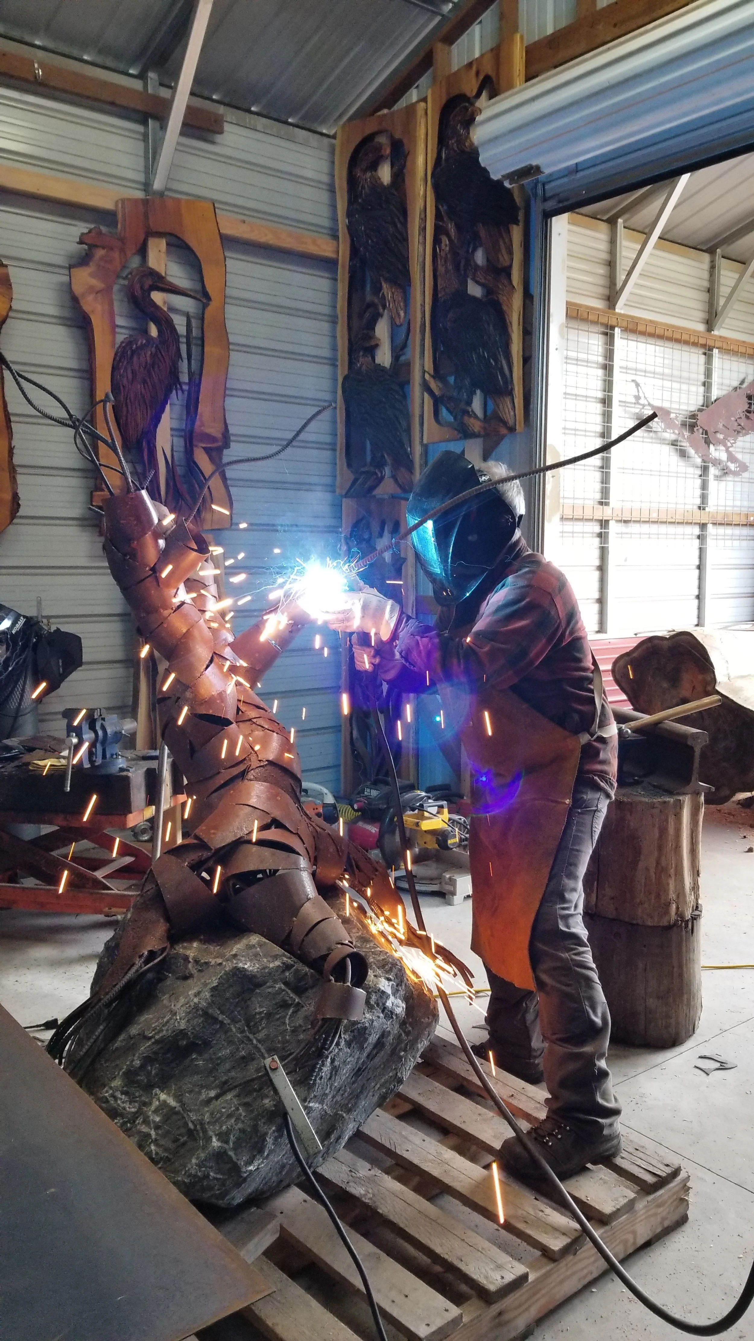 Bellingham sculptor Tomas Vrba welding a sculpture of a tree made of metal in his workshop. The workshop has wood and metal art pieces, including herons on the wall.