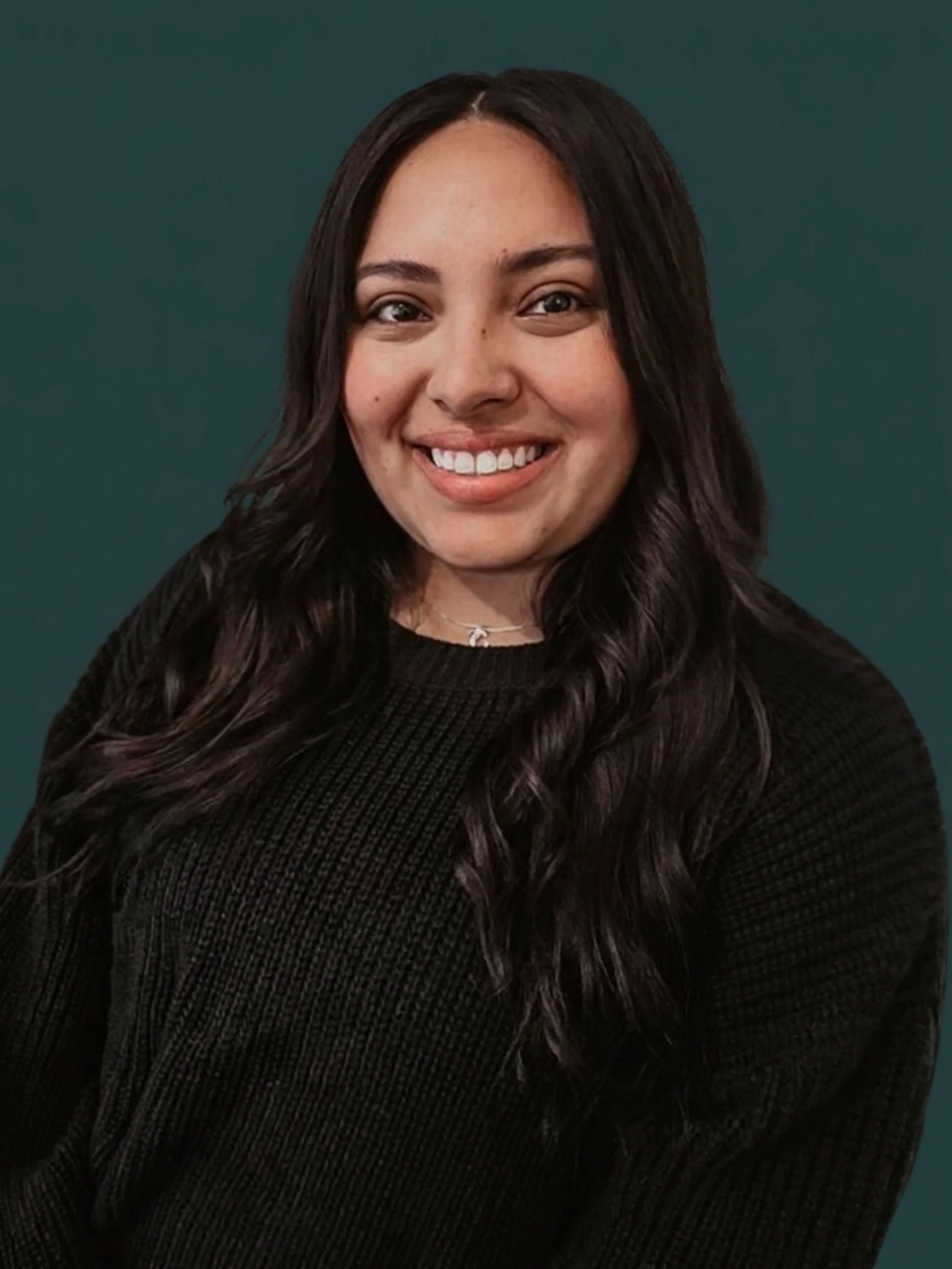 A smiling woman with long, dark, wavy hair wearing a black sweater and a silver necklace, standing in front of a dark green wall.