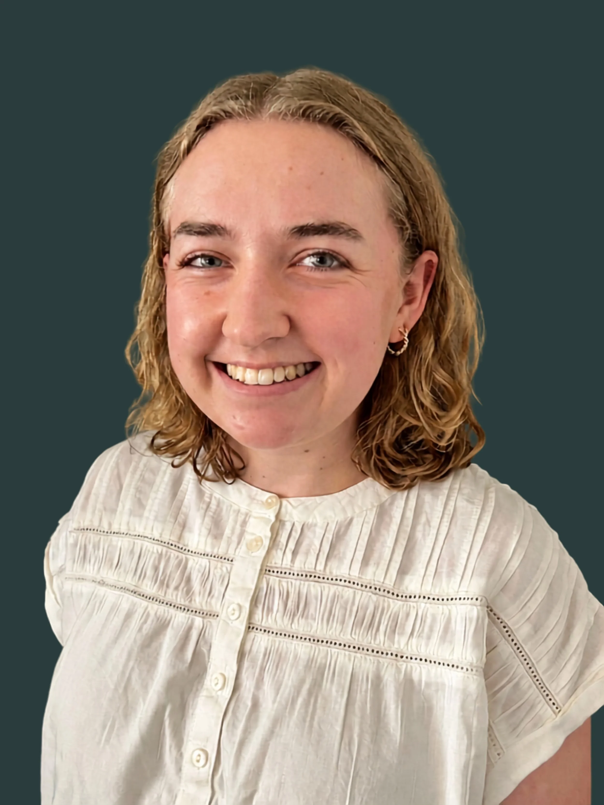 A woman with shoulder-length wavy hair, smiling, wearing a light-colored, button-up blouse with embroidery details, standing against a dark green wall.