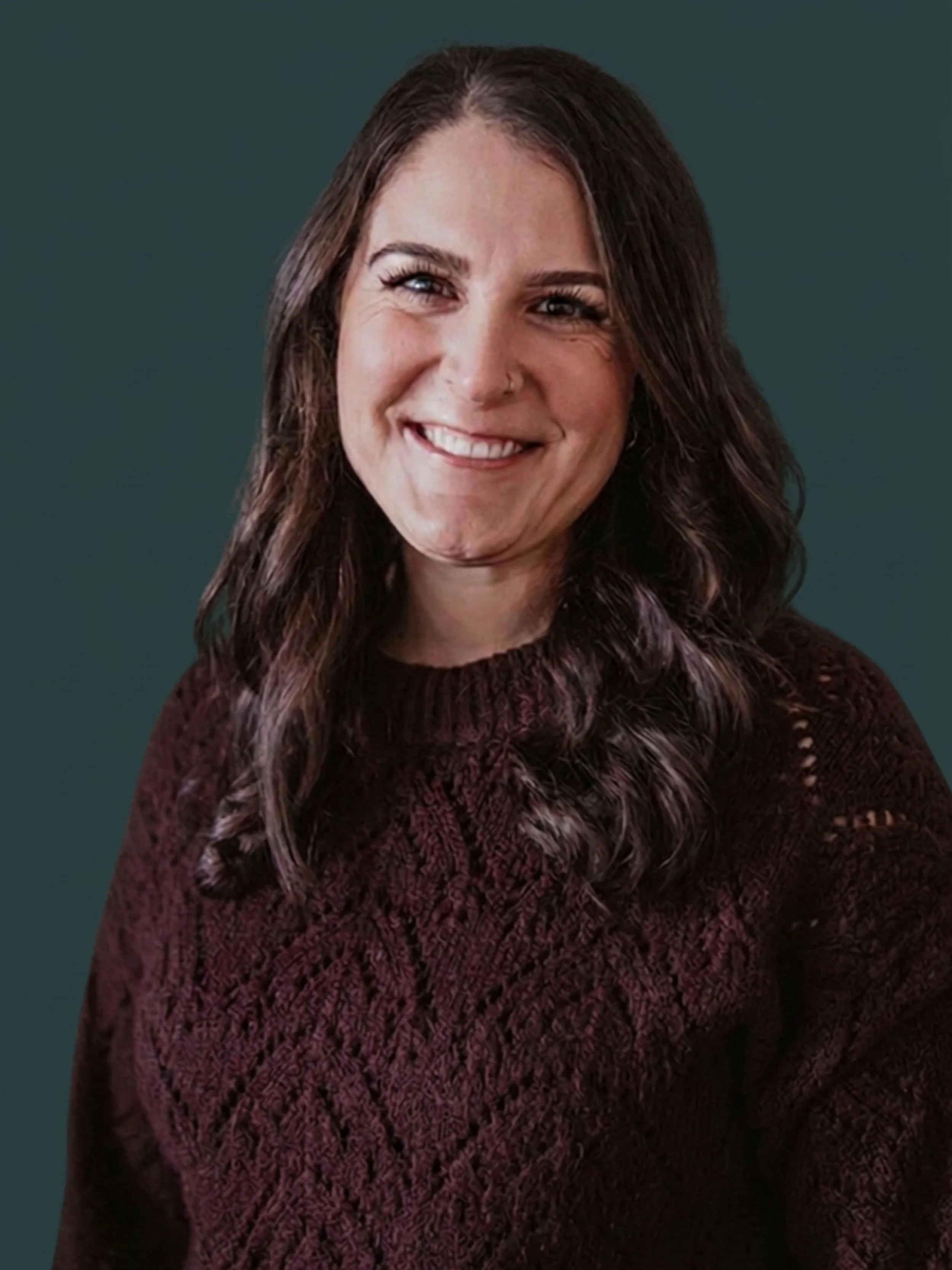 A woman with long dark wavy hair, wearing a maroon knit sweater, smiling in front of a background 