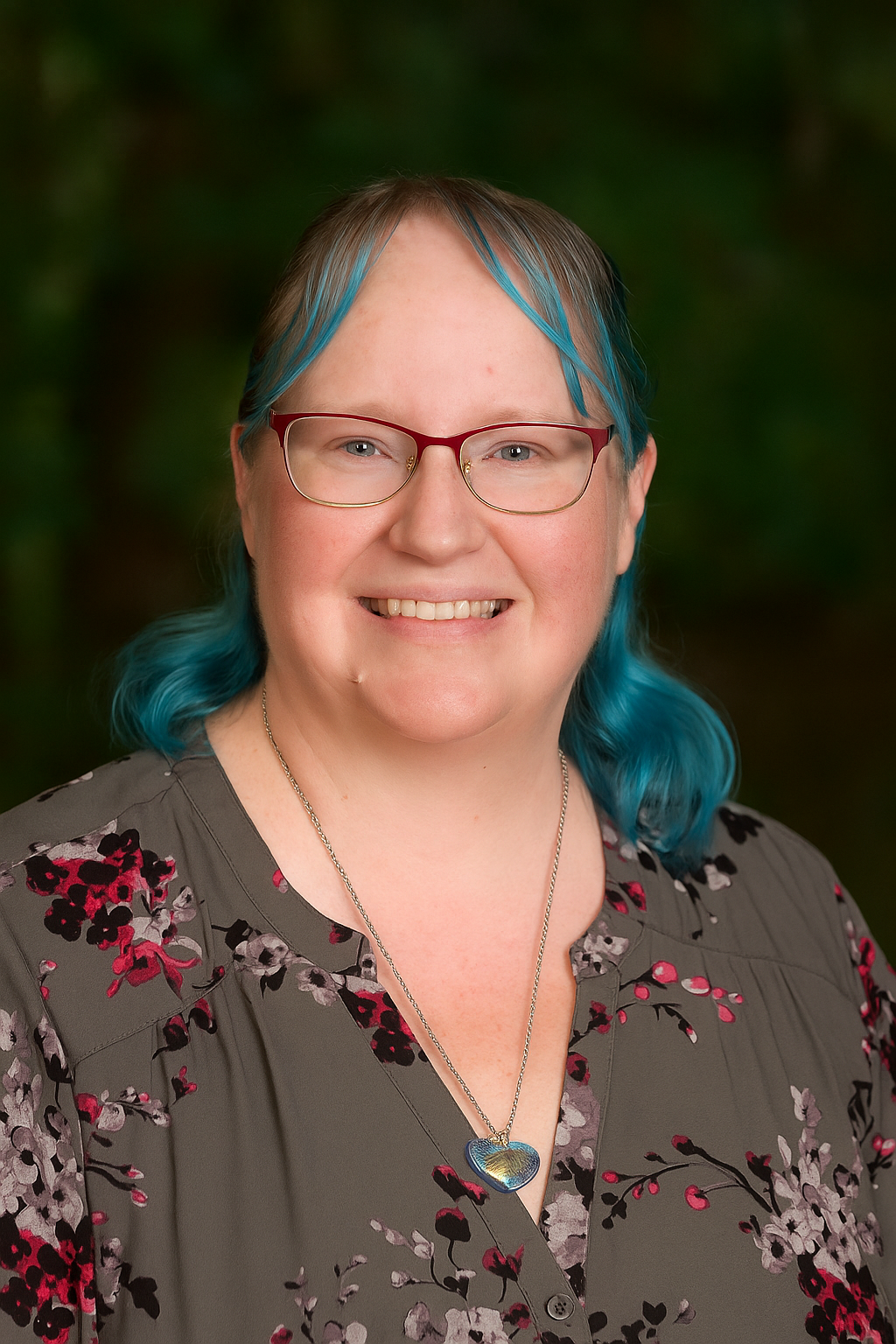 A woman with teal hair, glasses, and a floral blouse, smiling outdoors.