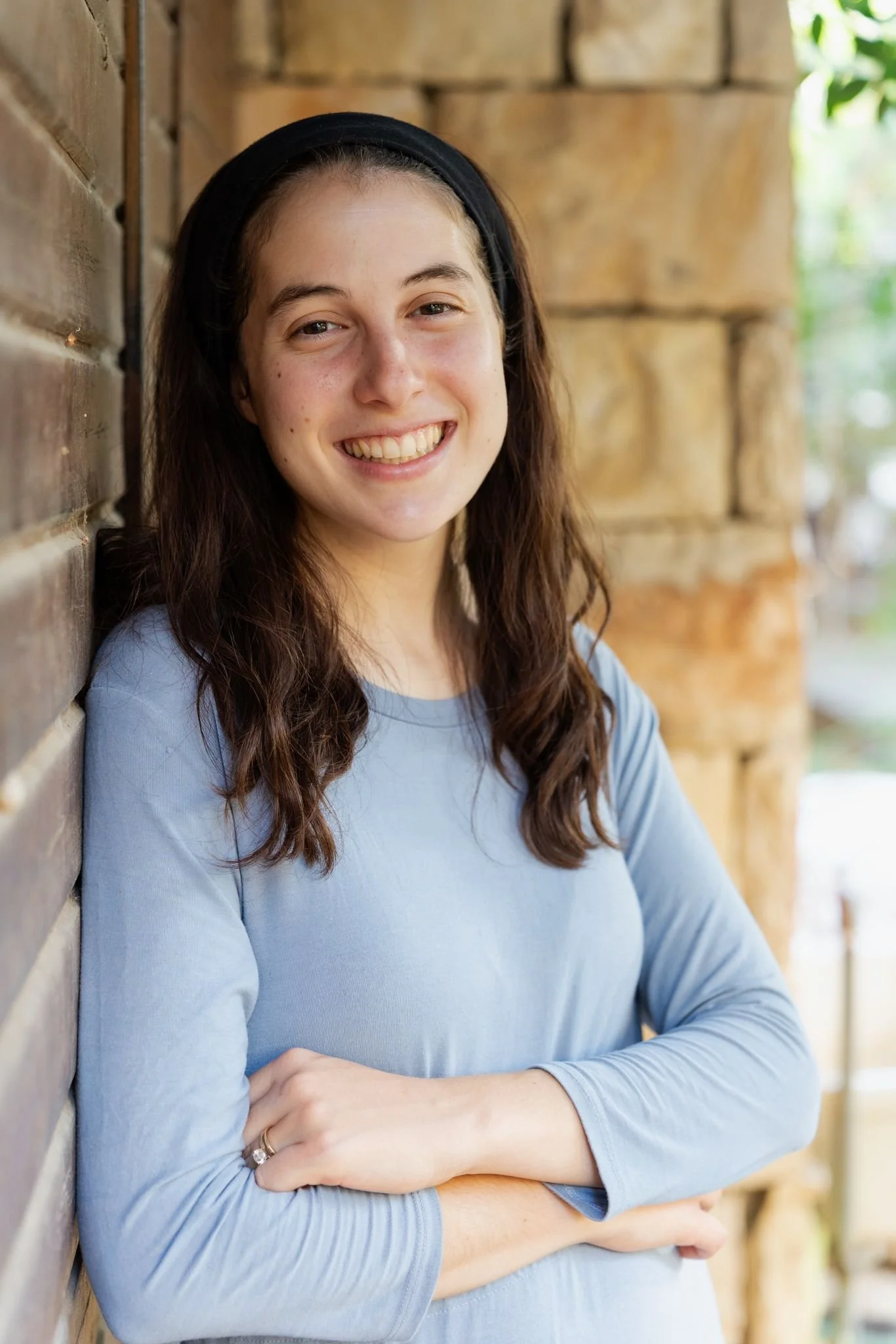 A young woman with long brown hair, smiling, wearing a black headband and a light blue long-sleeve shirt, standing next to a wooden wall outdoors.