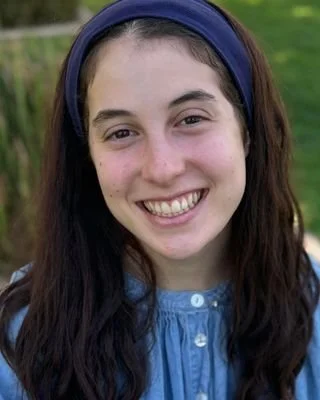 A smiling young woman with long dark hair wearing a blue headband and blue top, outdoors with green grass in the background.