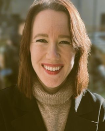 Close-up of a young woman with shoulder-length reddish-brown hair, smiling outdoors with sunlight, wearing a beige turtleneck and dark jacket.