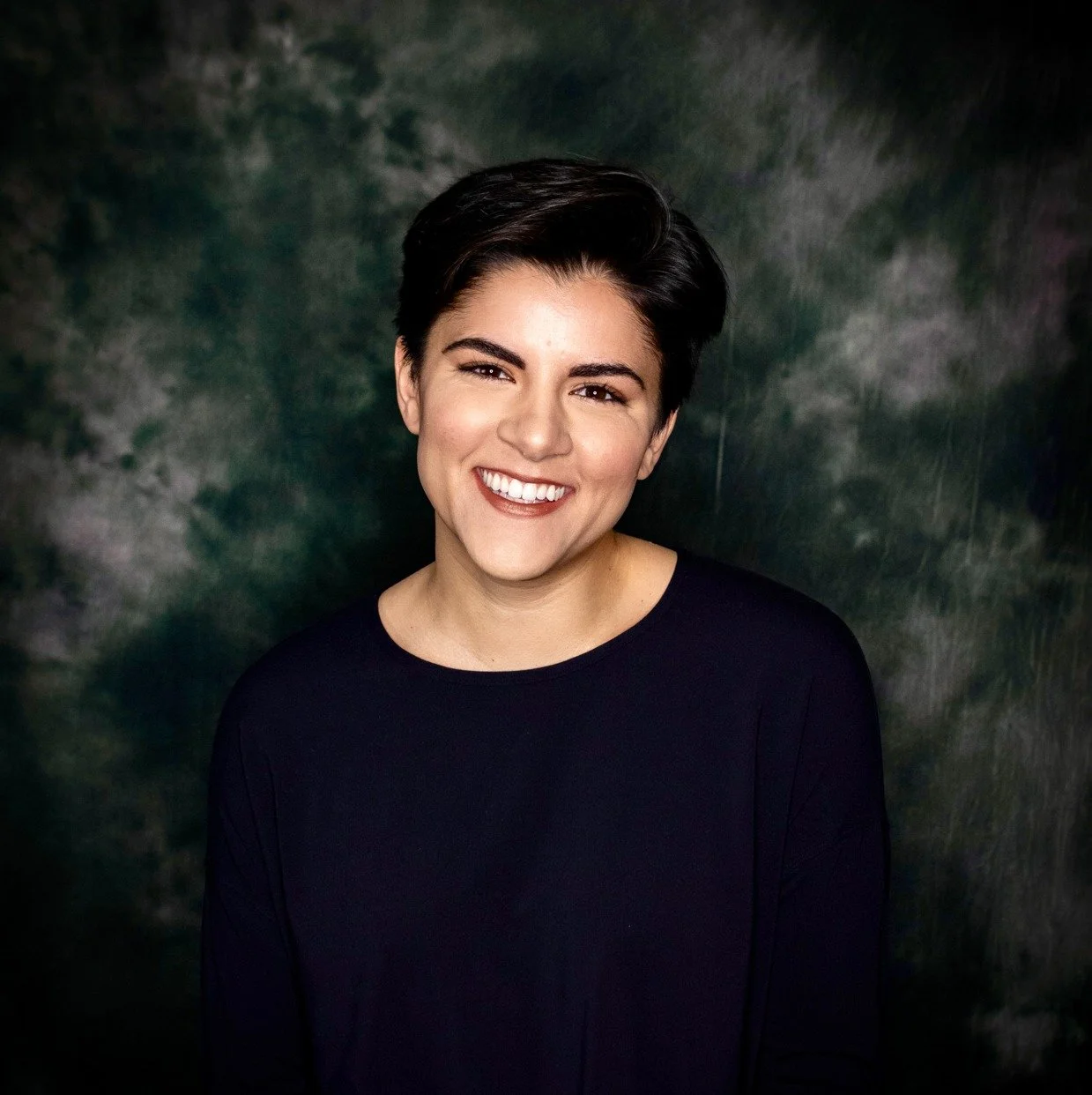 Portrait of a young woman with short dark hair, smiling, wearing a black top, against a green and gray mottled background.