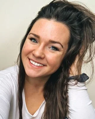 A smiling woman with long dark hair, wearing a white top, and holding her hair back with one hand.