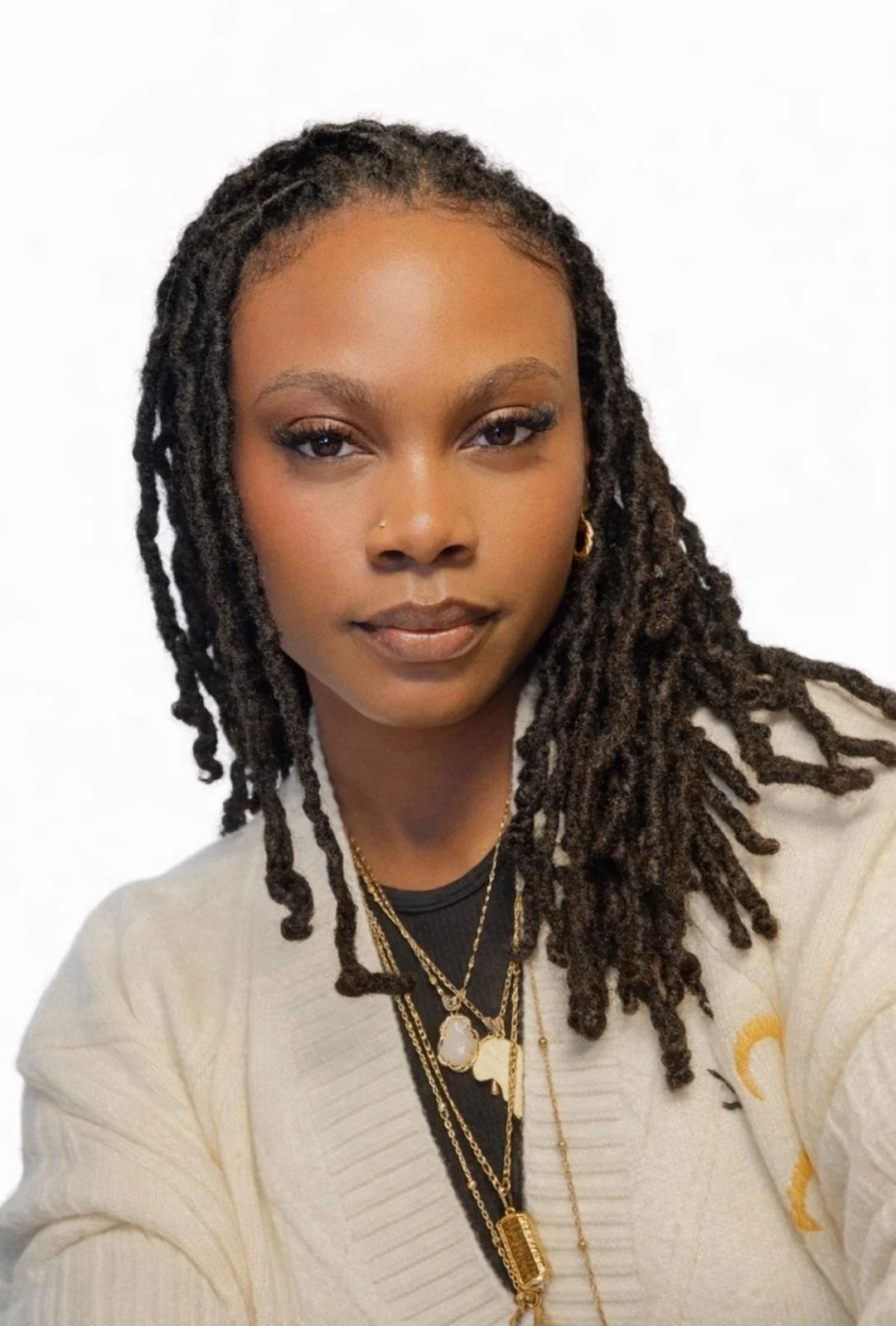 A woman with medium-brown skin, dreadlocked hair, and gold jewelry, including necklaces and earrings, looks confidently at the camera against a plain white background.