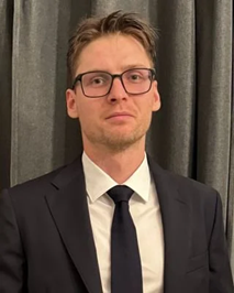 A man in a black suit, white shirt, and black tie standing in front of dark curtains.