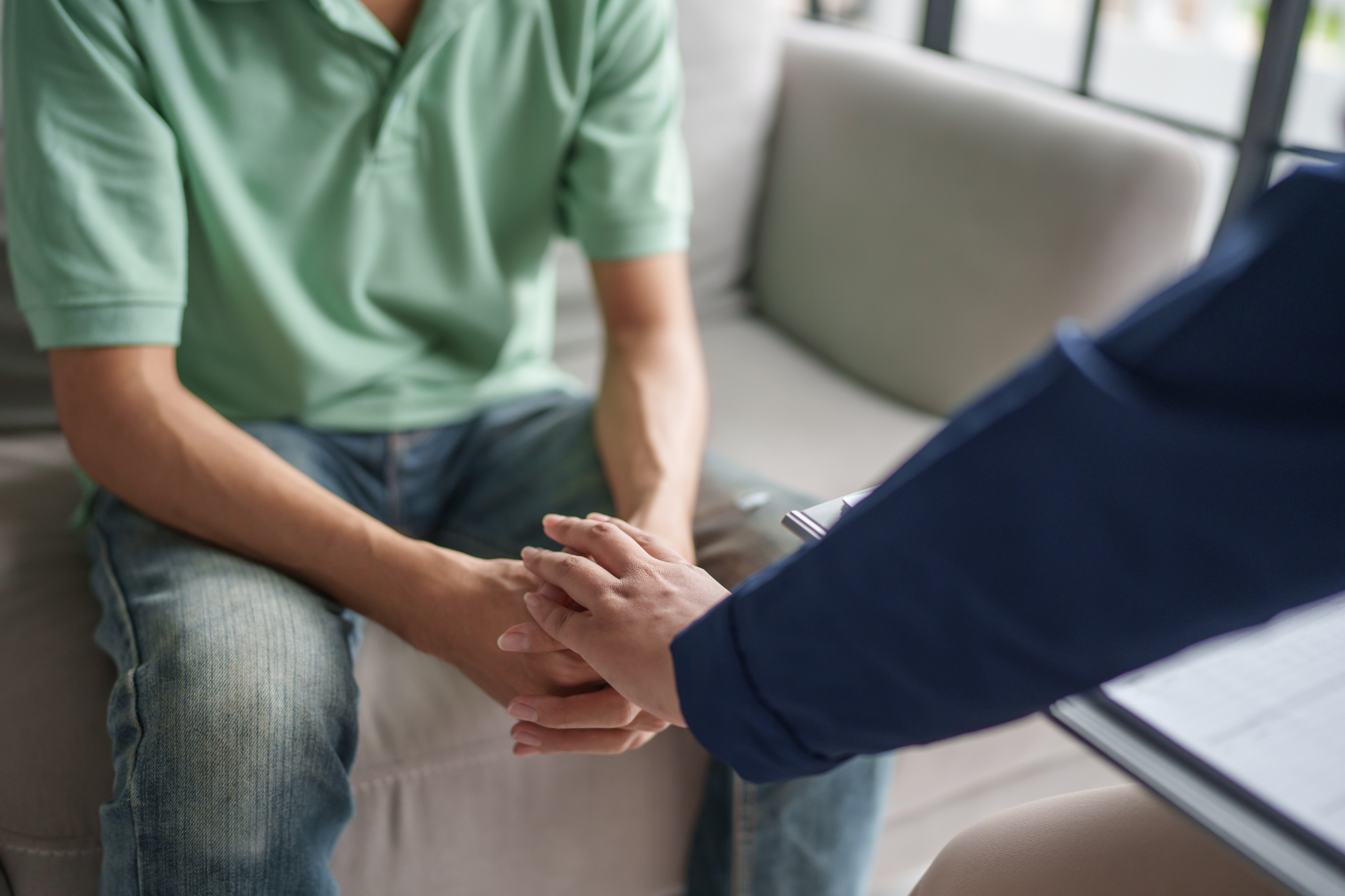 A person in a green shirt sitting on a sofa holding hands with another person in a blue shirt, during what appears to be a therapy or counseling session.