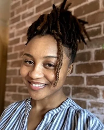 A woman with short dreadlocks smiling, wearing a blue striped top, standing in front of a brick wall.