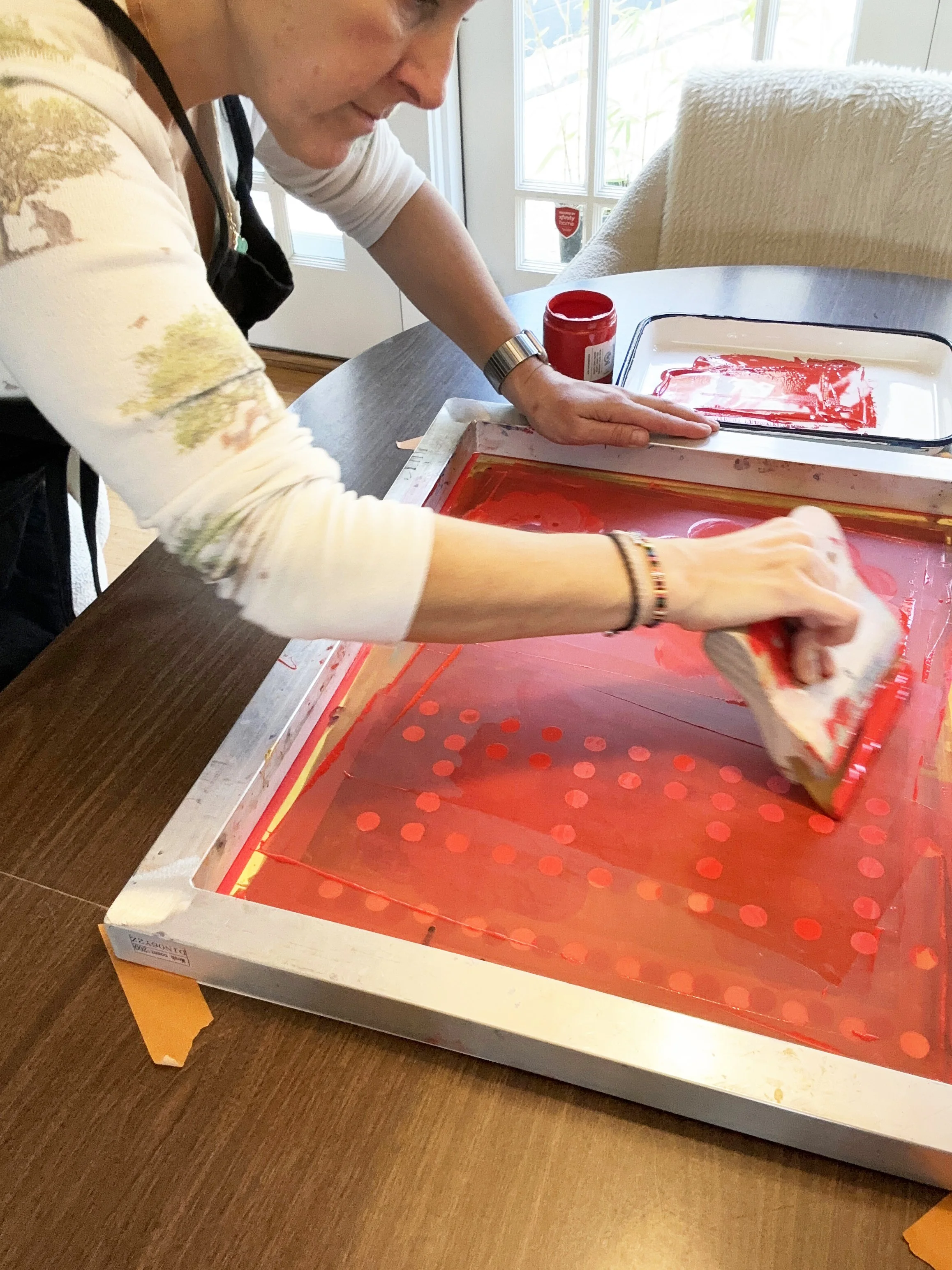 A person uses a squeegee to spread red paint or ink over a screen on a table, likely creating a print.