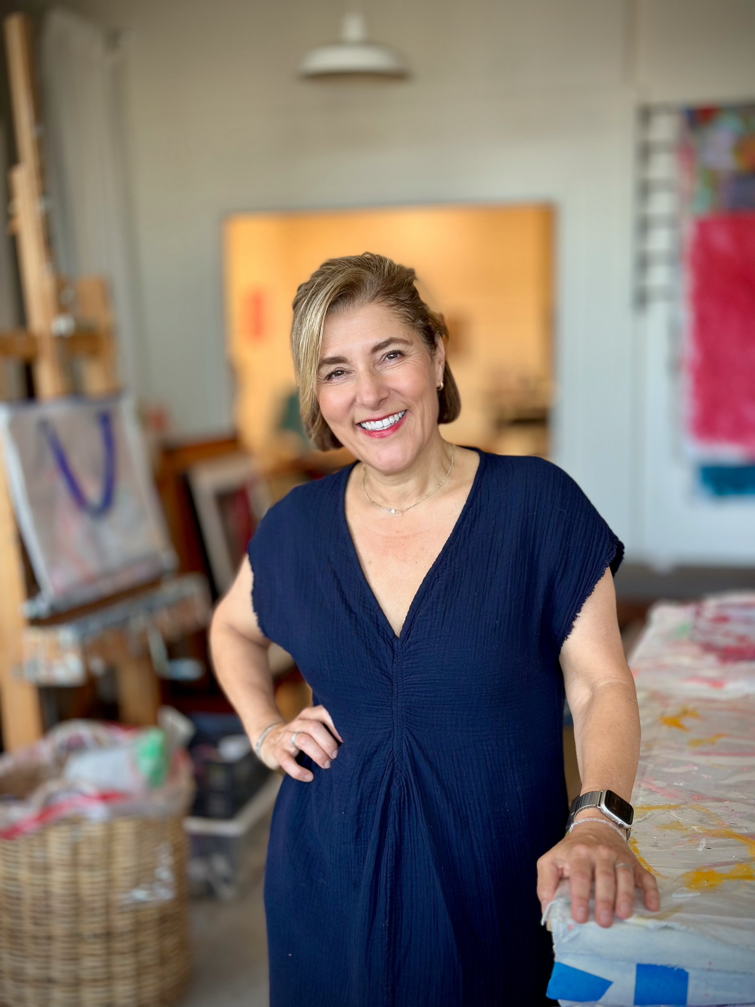 A smiling woman with short hair, wearing a navy blue dress, stands in an art or craft studio with shelves of art supplies and baskets in the background.