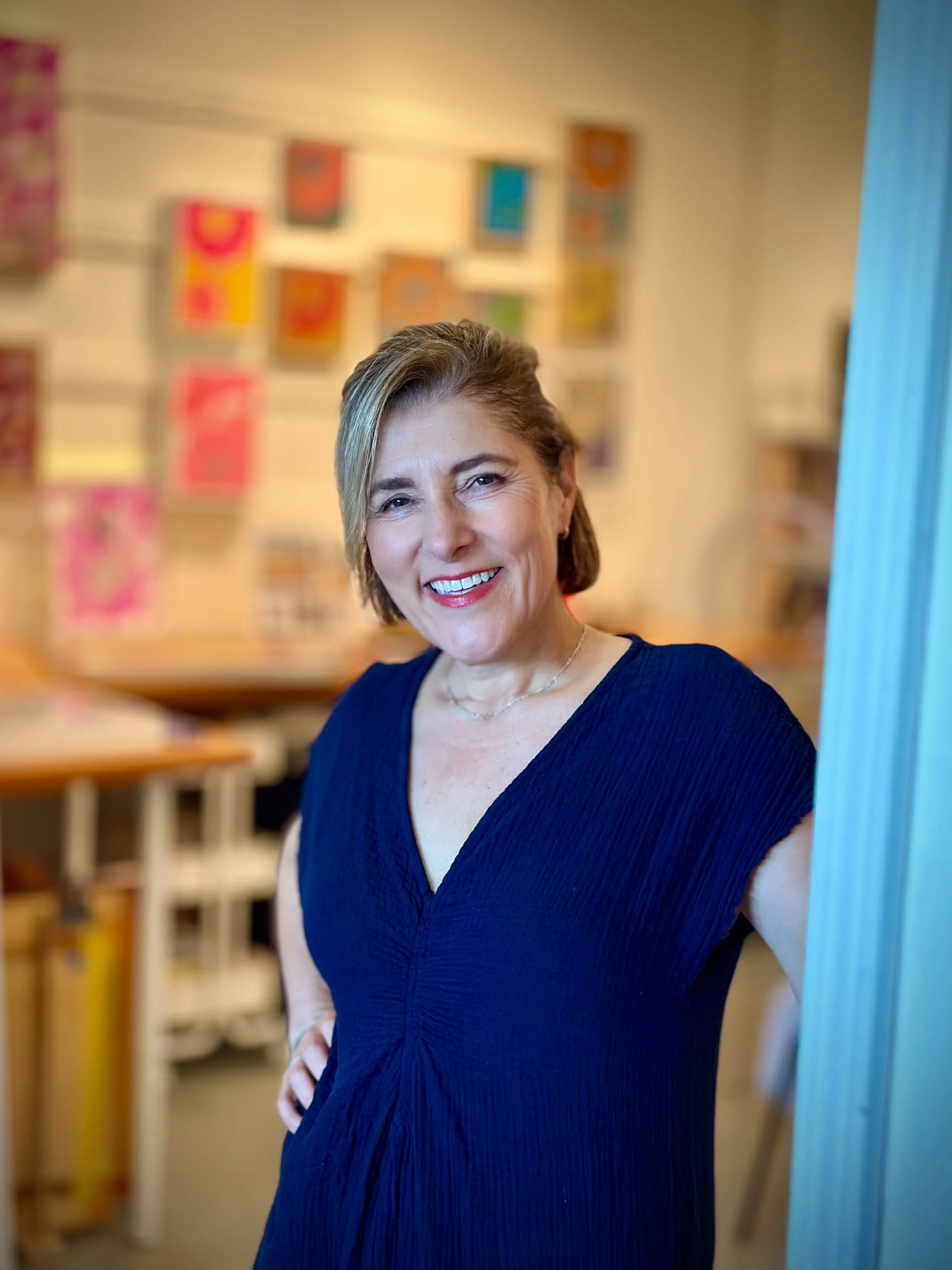 A smiling middle-aged woman with shoulder-length brown hair wearing a navy blue dress, standing indoors with colorful artwork on the wall behind her.