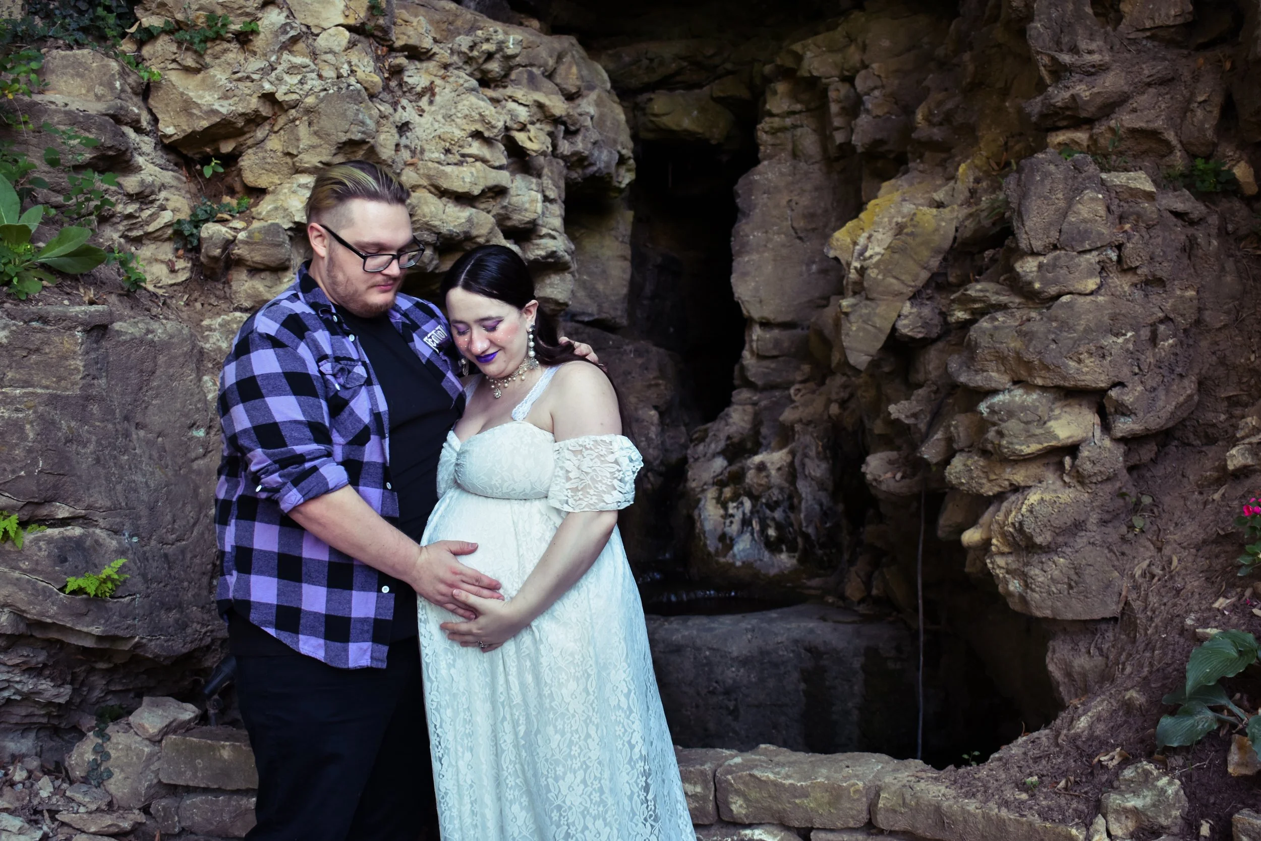 Pregnant couple embracing in front of rocky background, woman in white lace dress, man in plaid shirt.