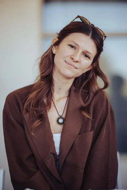A woman with wavy brown hair wearing a brown blazer and glasses on her head, smiling softly.