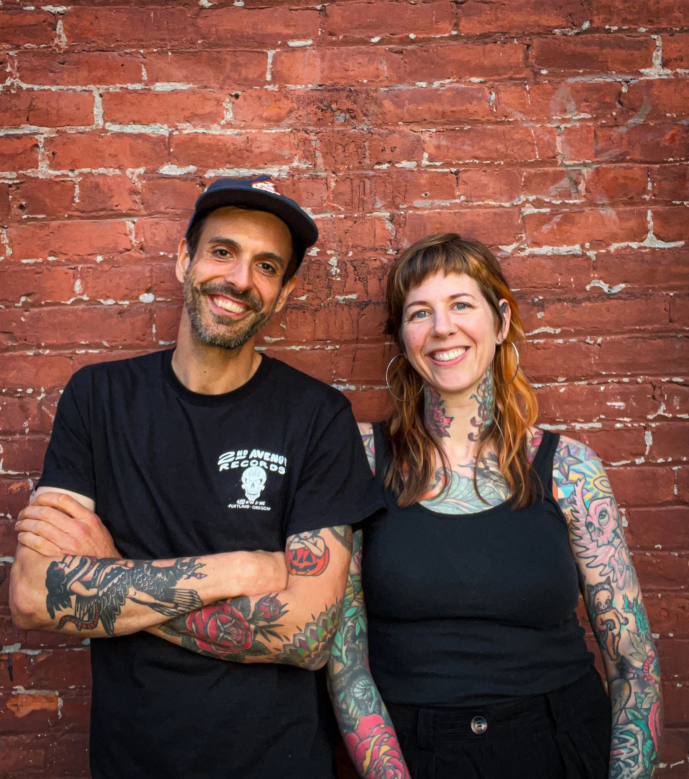 Two smiling individuals, a man wearing a black cap and T-shirt with logo, and a woman with tattoos on arms and chest, standing in front of a red brick wall.