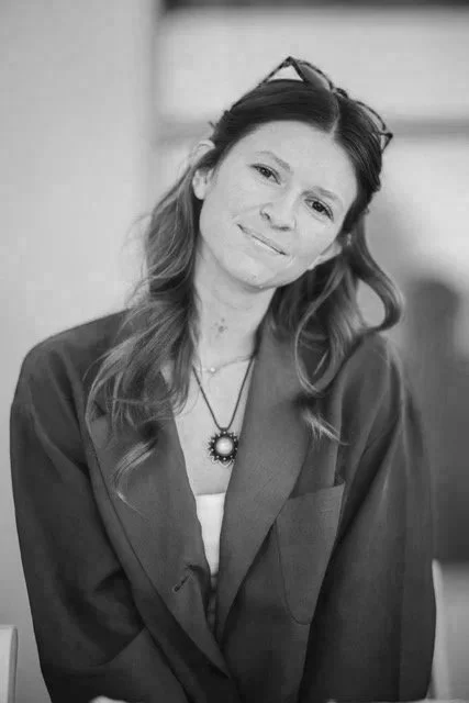 A woman with wavy brown hair wearing a brown blazer and glasses on her head, smiling softly.