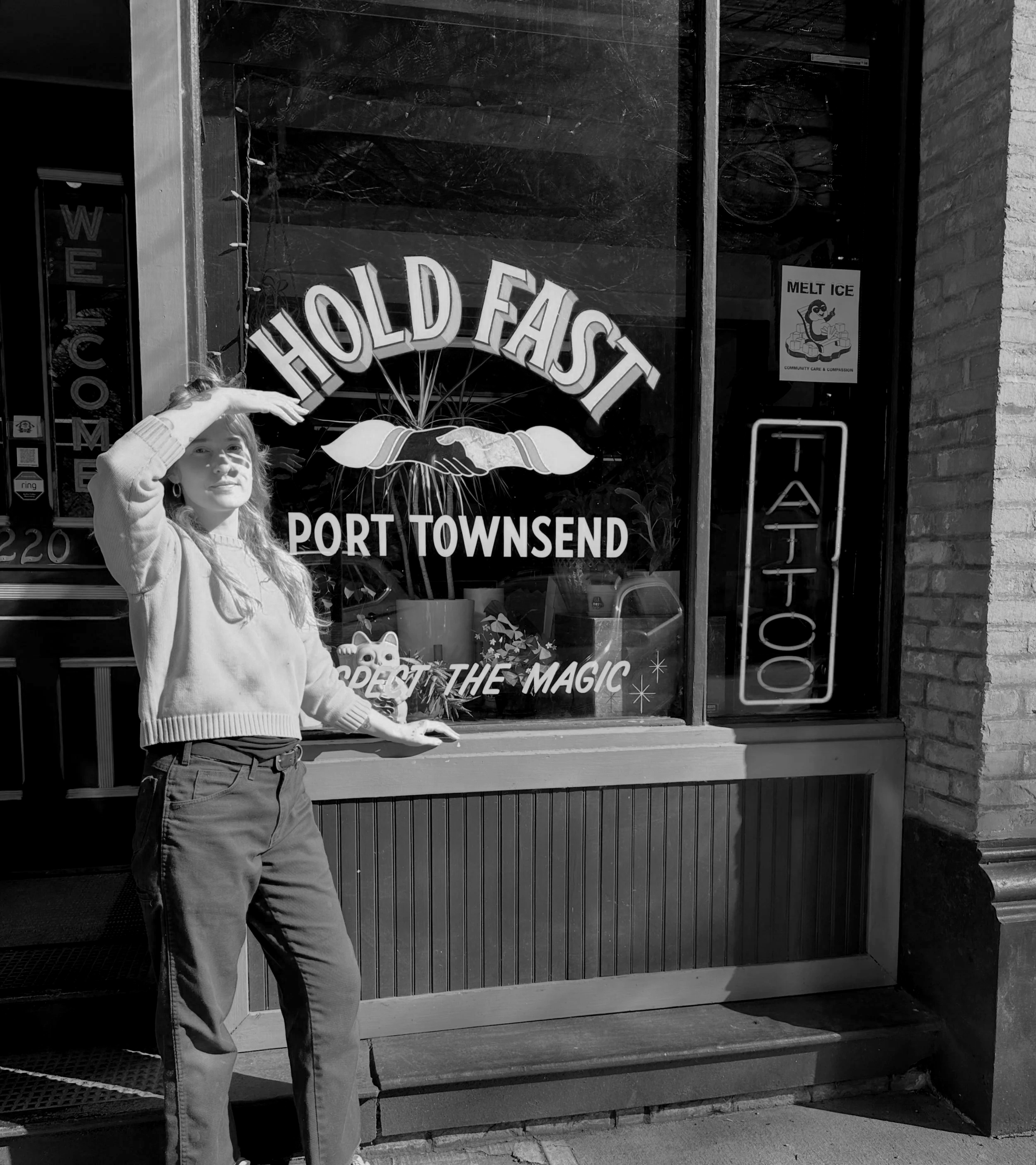 A young woman with red hair saluting in front of a glass storefront window advertising an ice cream shop named "Hold Fast" in Port Townsend. The window displays a yellow poster with a cartoon ice cream sitting on ice, labeled "Melt Ice." The woman is wearing a beige sweater and blue jeans, and there is a small MANEKI NEKO or lucky cat figure on the window ledge.