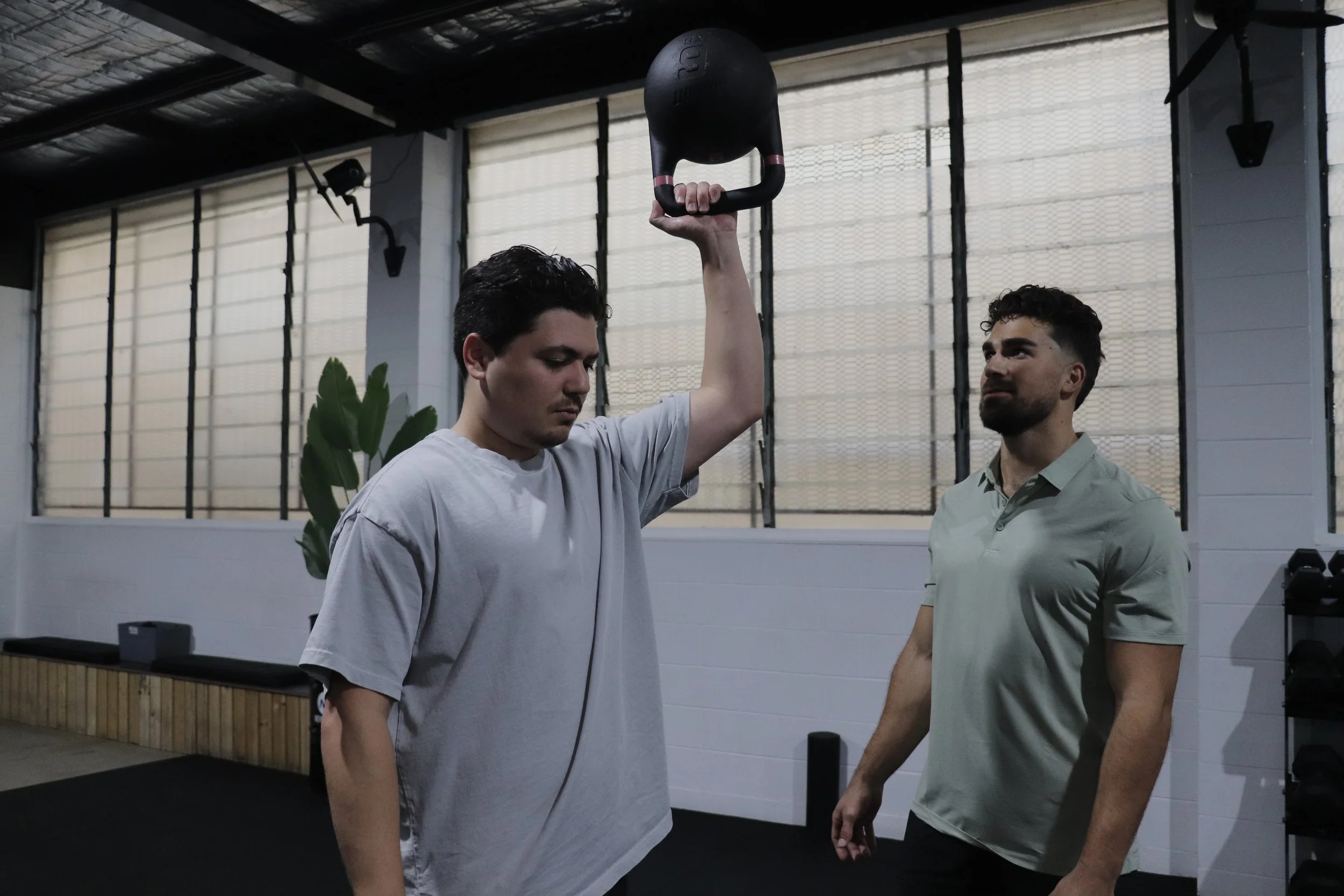 Man holding a kettlebell overhead with a trainer observing in a gym.