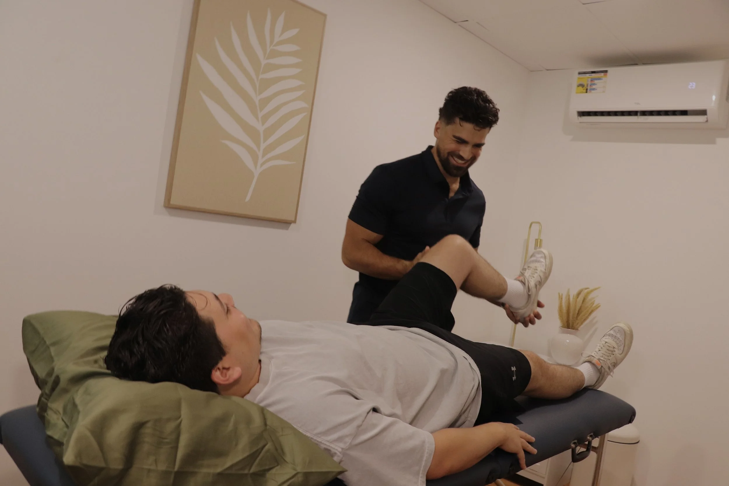 A man lying on a massage table being assisted by a chiropractor or therapist, who is holding his legs up. The man appears relaxed and is looking at the ceiling, while the therapist is smiling. The room has minimal decor with a beige and white color s