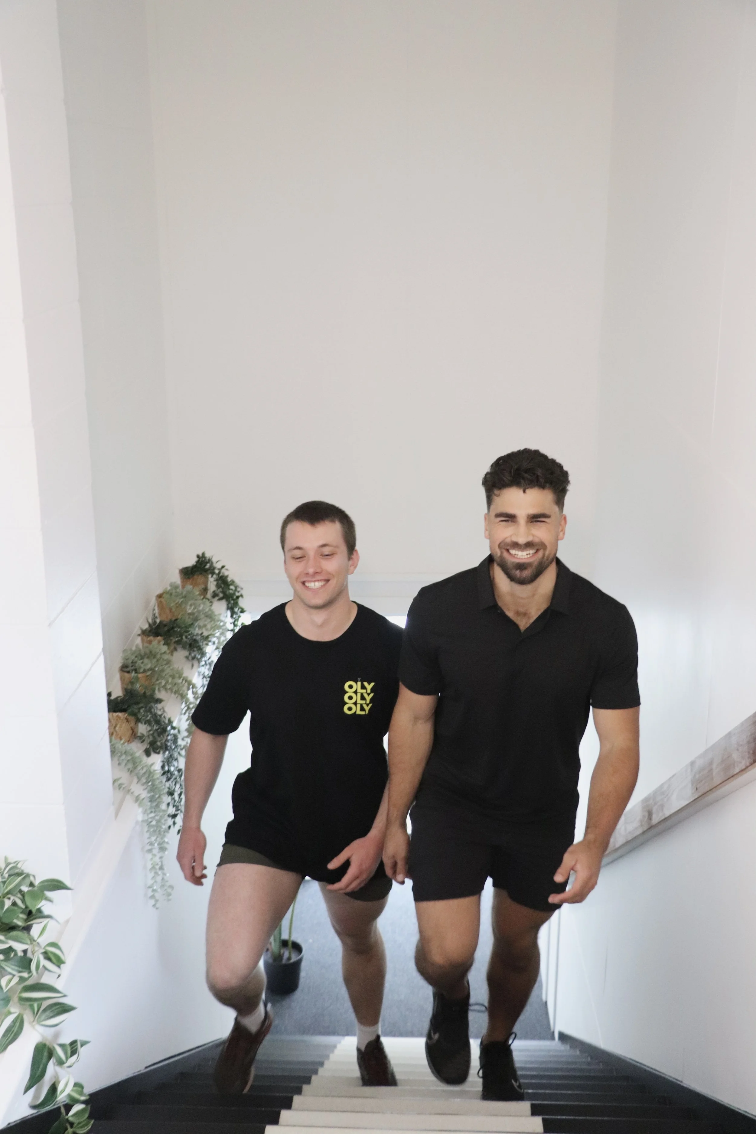 Two men walking up a staircase smiling, one wearing a black t-shirt with yellow text and shorts, the other in a black polo shirt and shorts, with plants on the side of the staircase.