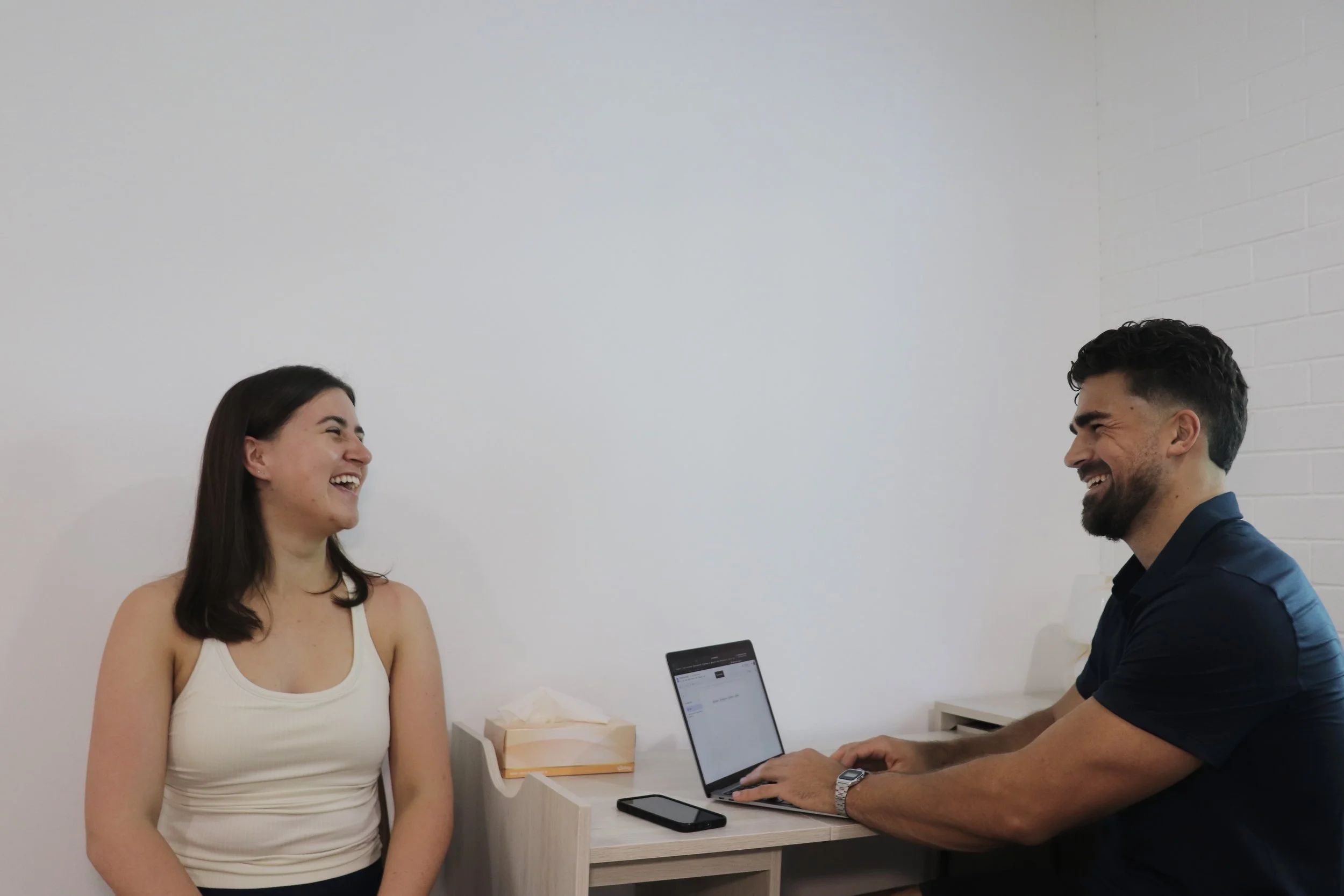 A woman and a man laughing and talking while sitting at a desk with a laptop, tissue box, and smartphone.