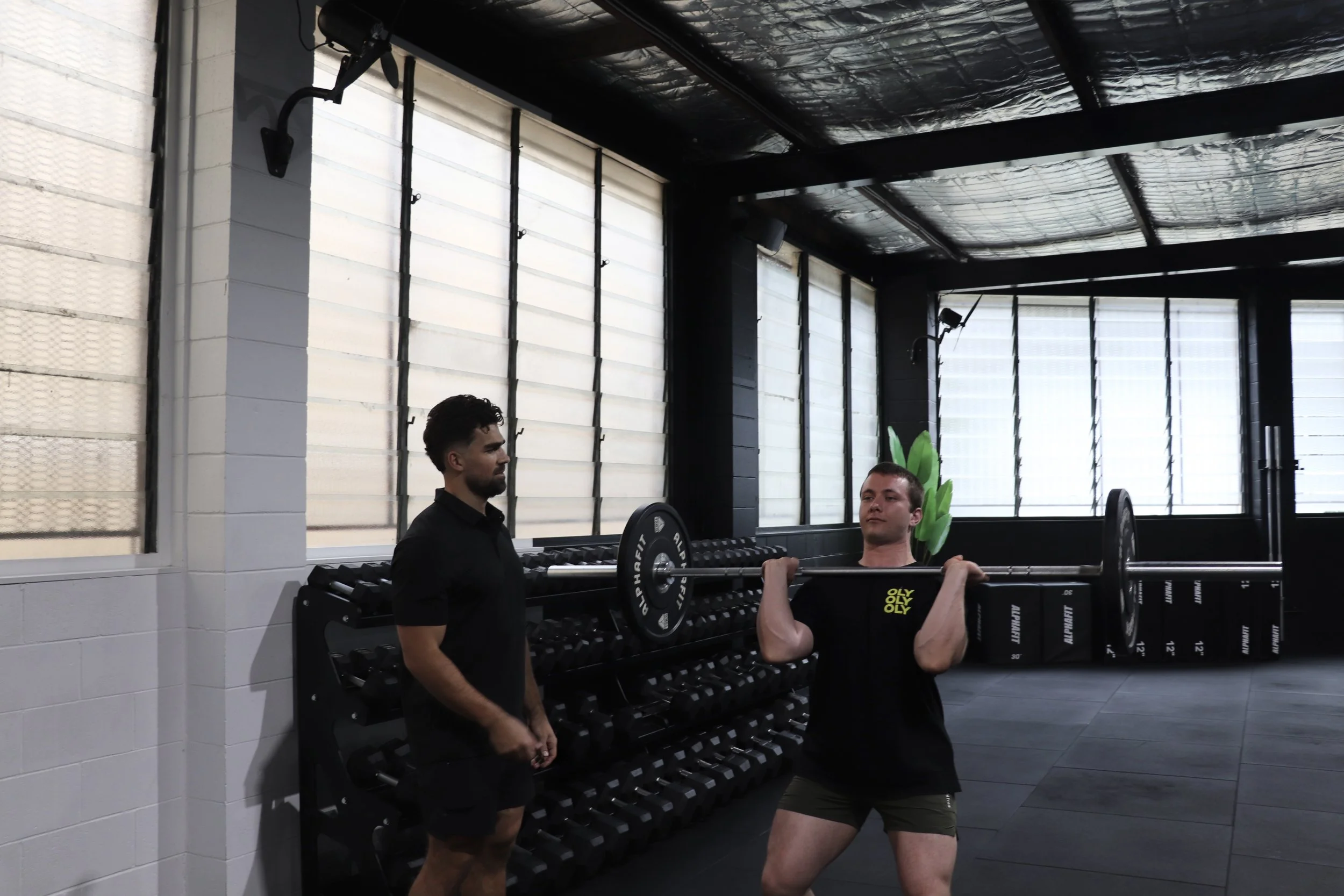 A man lifts a barbell in a gym while another man observes. The gym has large windows, black walls, and weight racks.