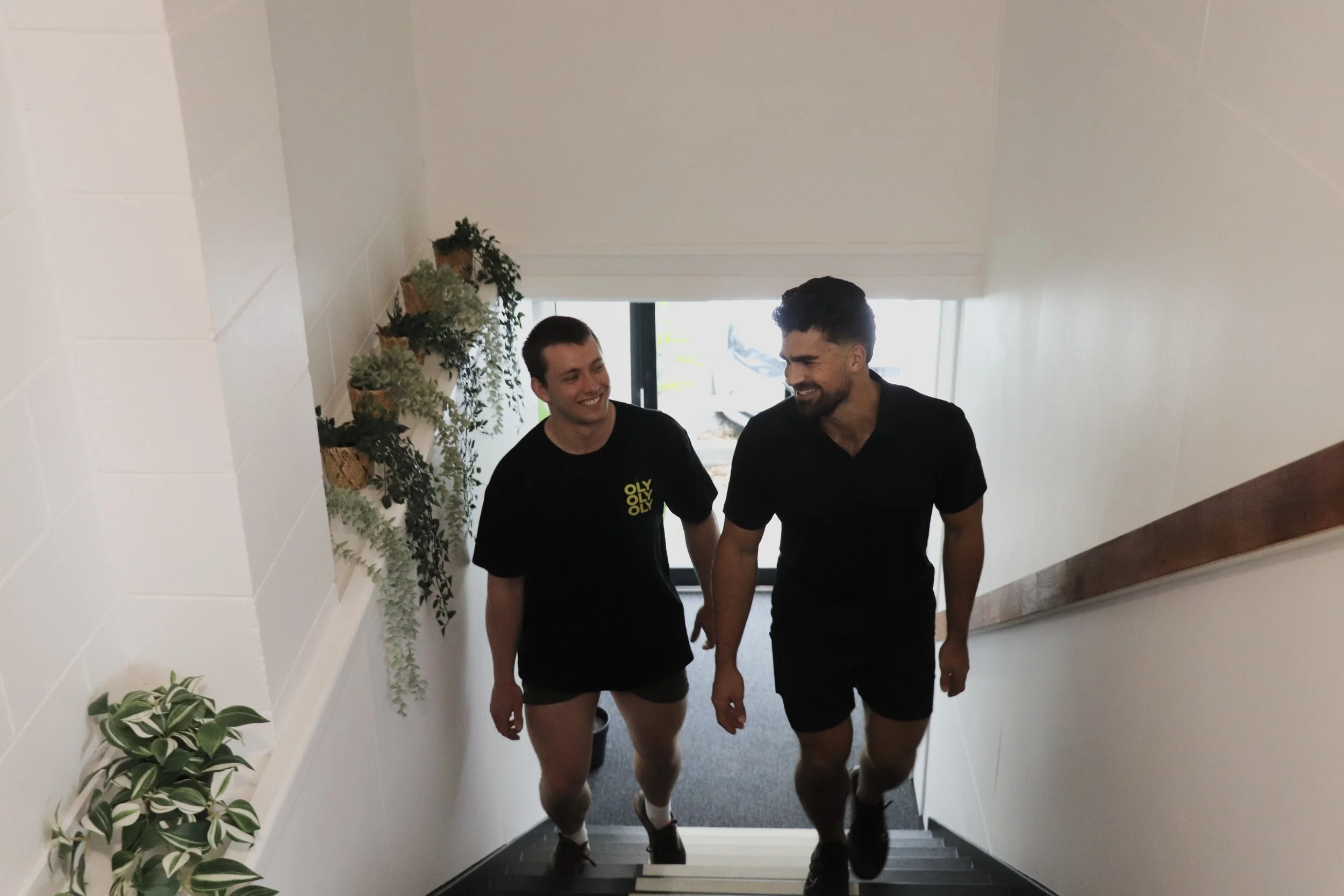 Two young men walking up stairs in a well-lit indoor space, smiling at each other. One is wearing a black shirt with yellow text, and the other is in a black polo shirt. They are holding hands.