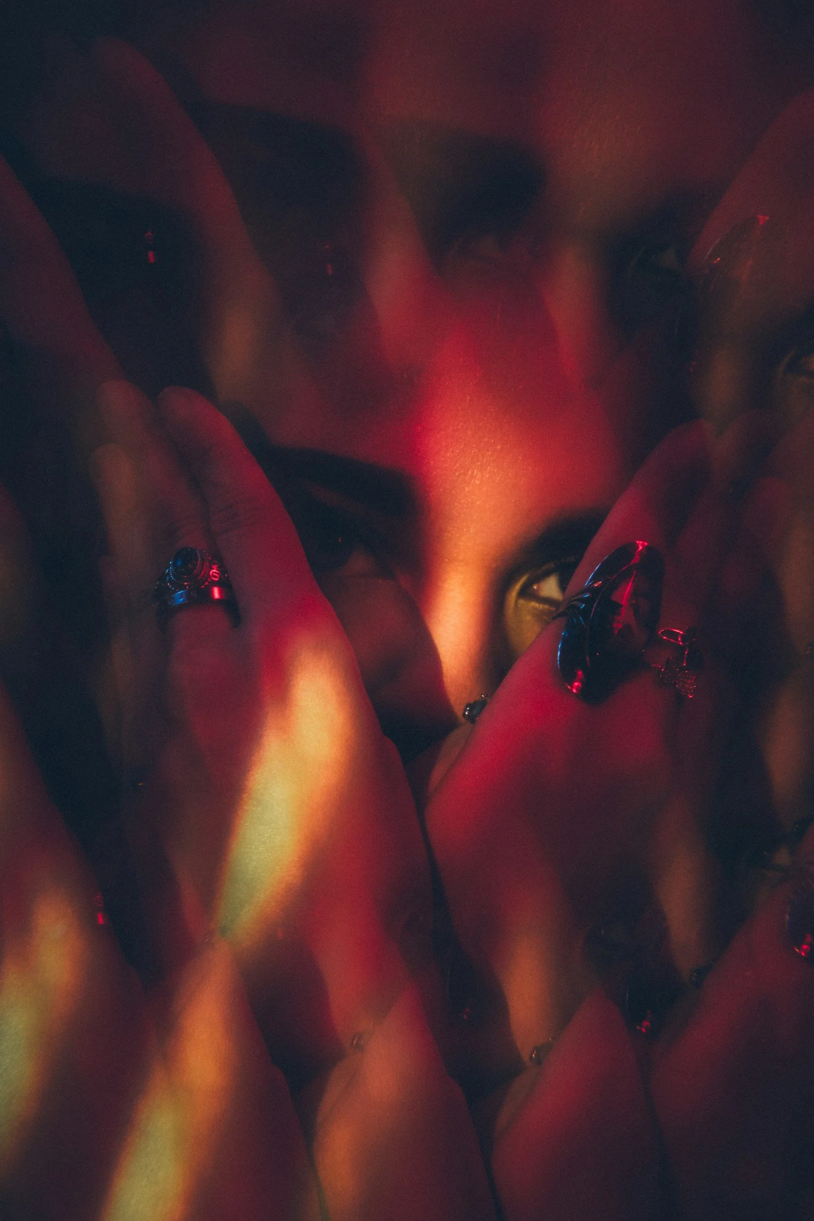 Multiple hands with rings overlapping and touching a person's face in a dimly lit environment with red lighting.